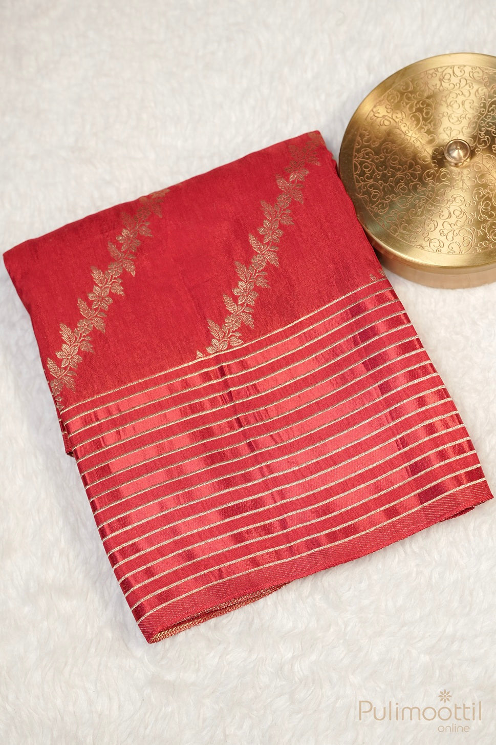 Red saree with gold floral and striped design, displayed with decorative gold container on white background