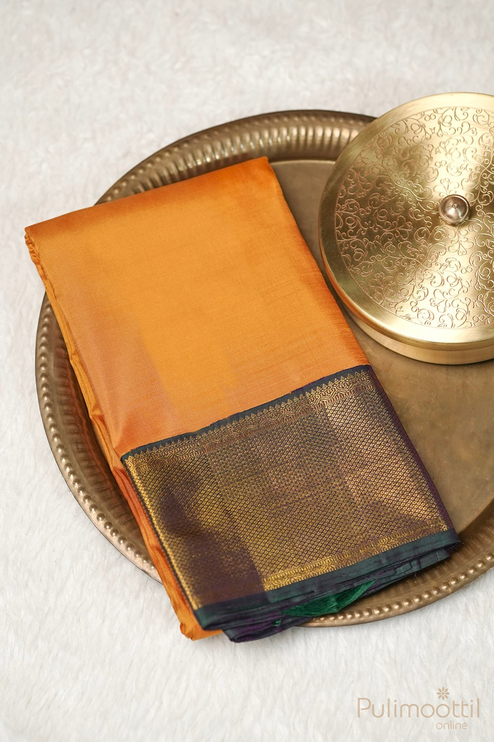 "Mustard yellow Kanchipuram soft silk saree with a plain body and a contrasting golden and purple border, elegantly placed on a decorative brass plate with an intricate lid beside it.