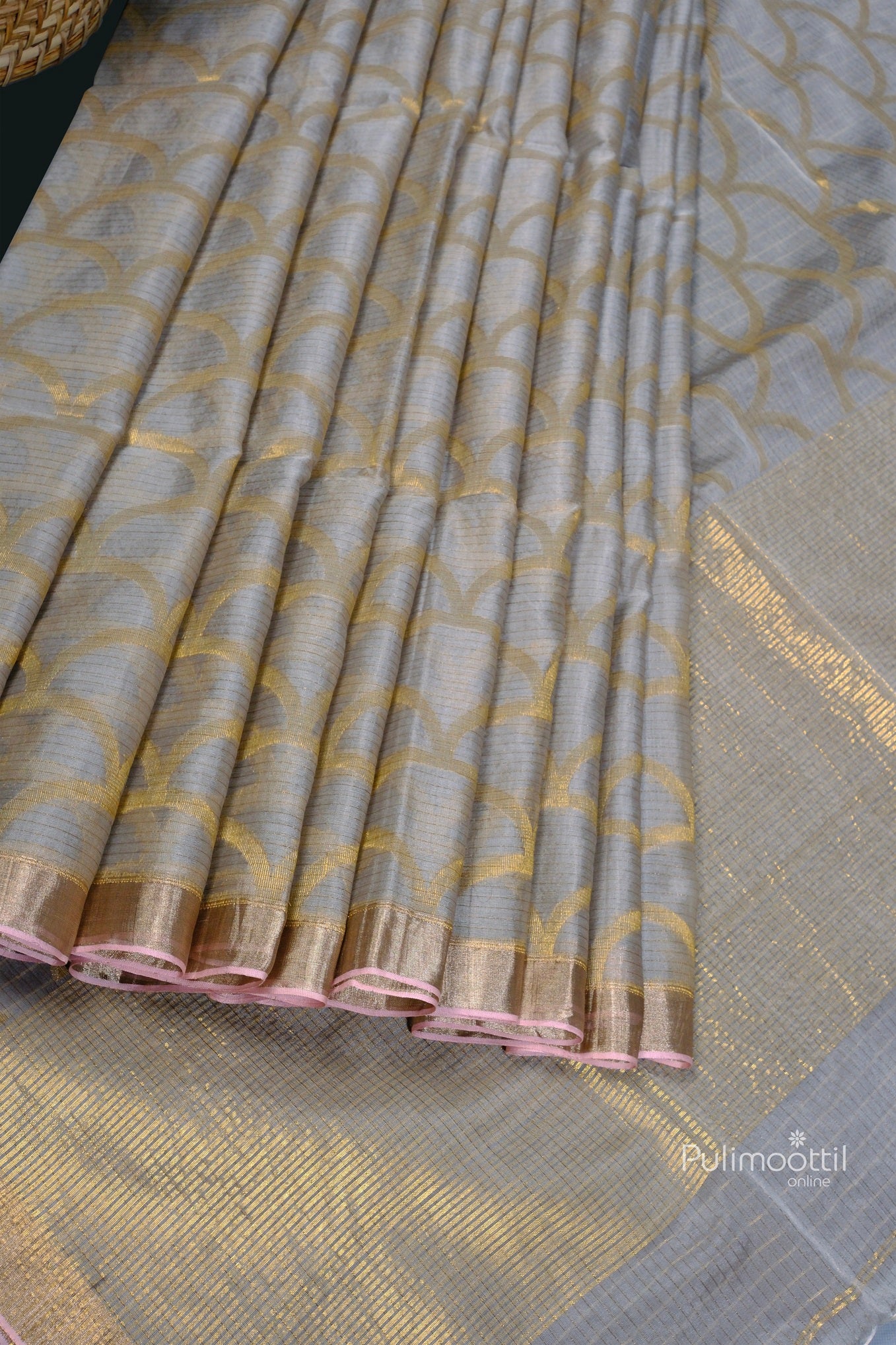 Close-up of pleated ash Tussar silk saree with intricate golden scallop patterns and a soft pink selvedge