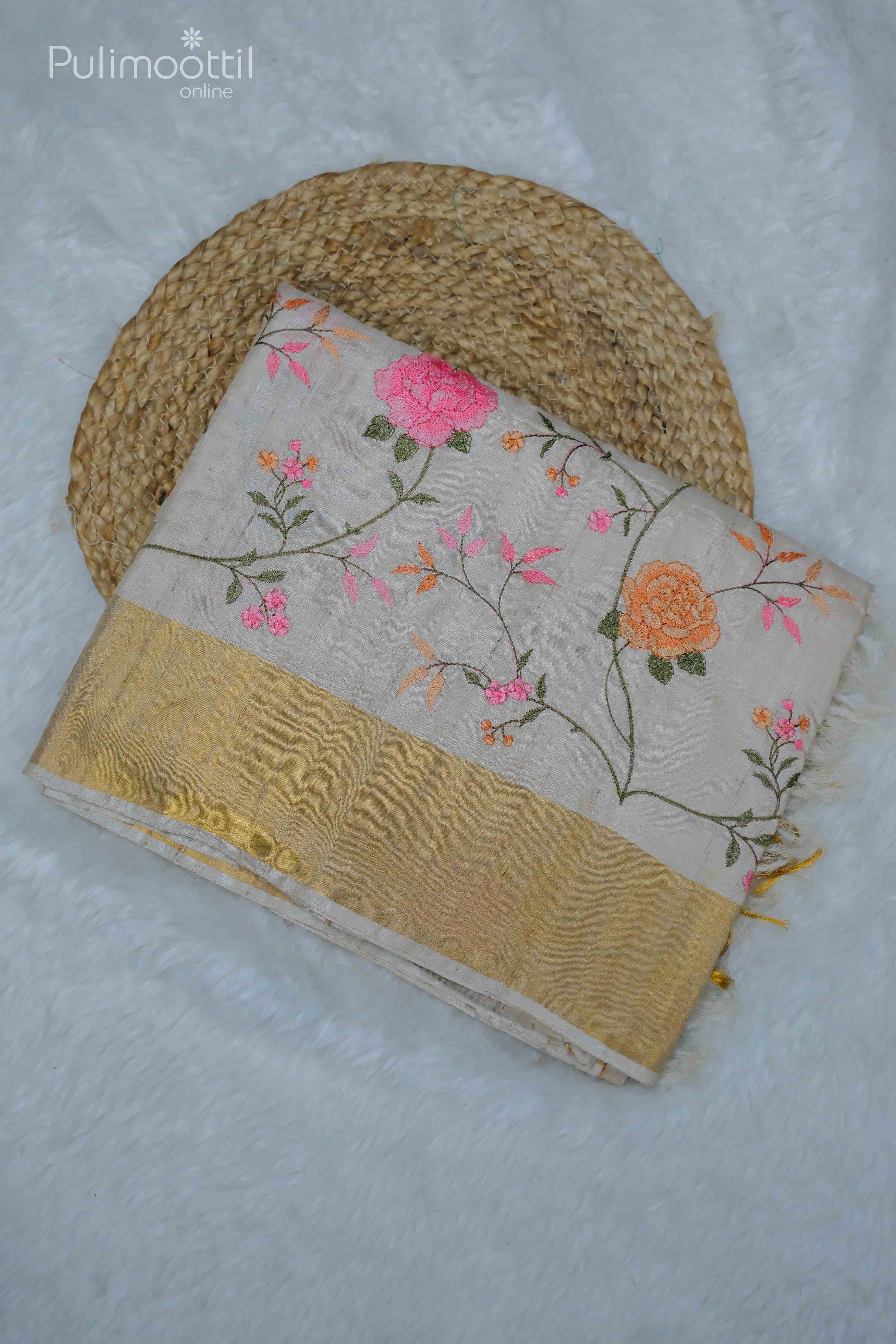 Beige pure tussar silk saree with floral embroidery in pink and orange hues, featuring a golden tissue border, neatly folded on a round woven mat.