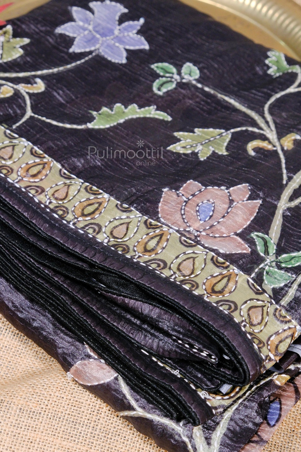 Folded black saree with purple and beige embroidered flowers and a paisley print border on beige fabric.