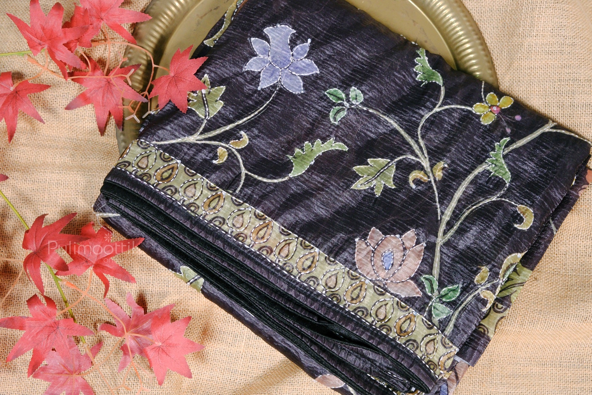 Full view of a folded black floral silk saree placed on a golden plate with decorative red leaves around it.