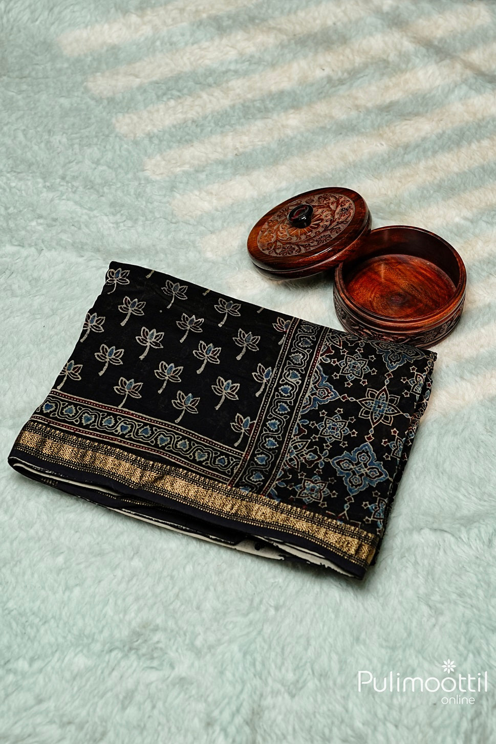 Folded Black Colour Chandheri Silk Saree with floral block print and zari zig-zag border, accompanied by a wooden storage box.