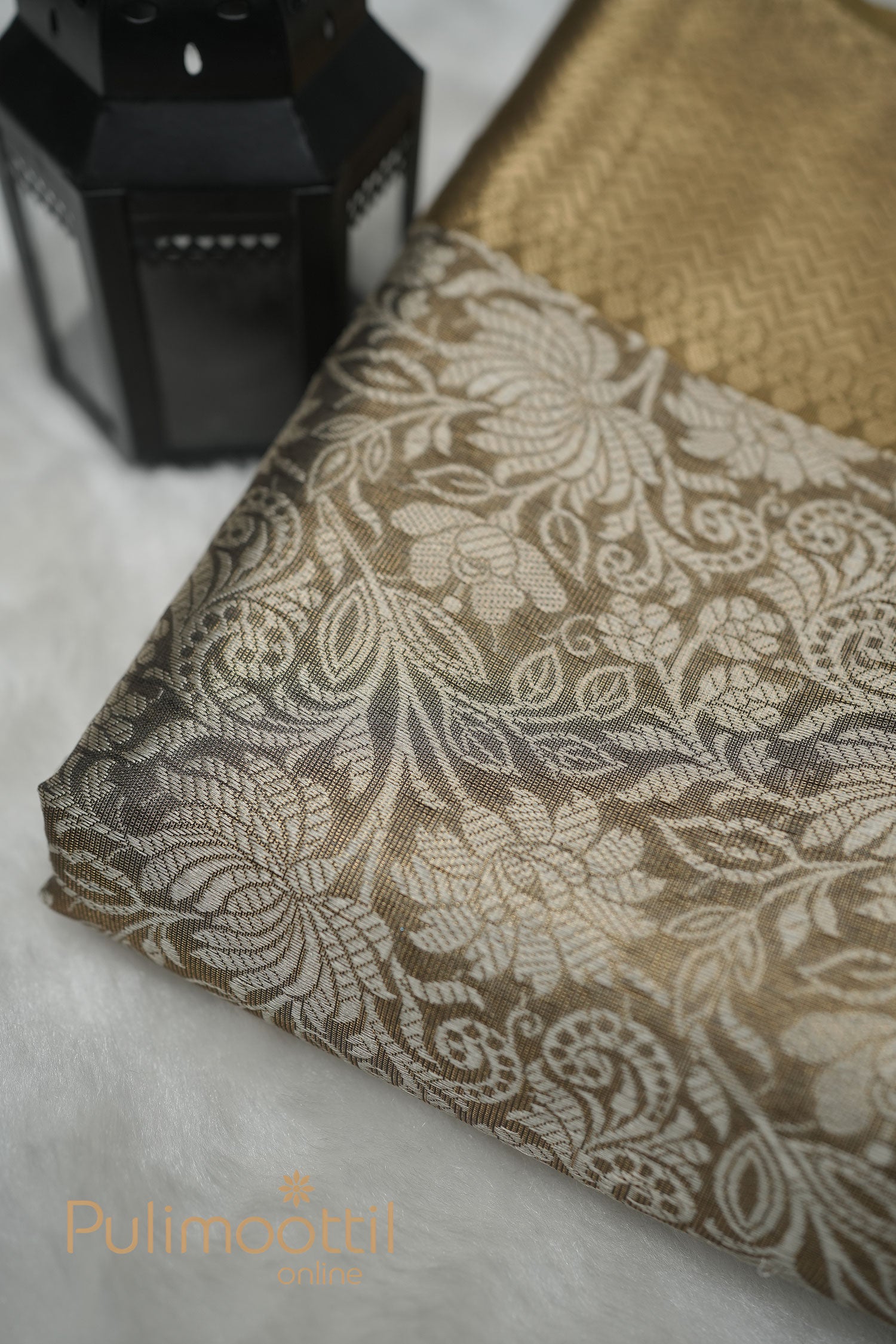 Gold‑beige semi‑silk fabric with intricate floral and paisley woven motifs draped beside a black lantern on a white plush surface.
