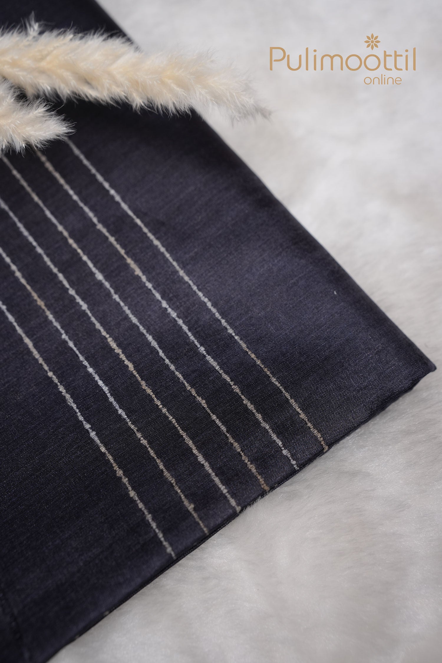 Folded charcoal black Tussar silk saree with fine vertical self-thread lines, placed on a soft white background with fluffy beige grass decor – Pulimoottil branding on top right
