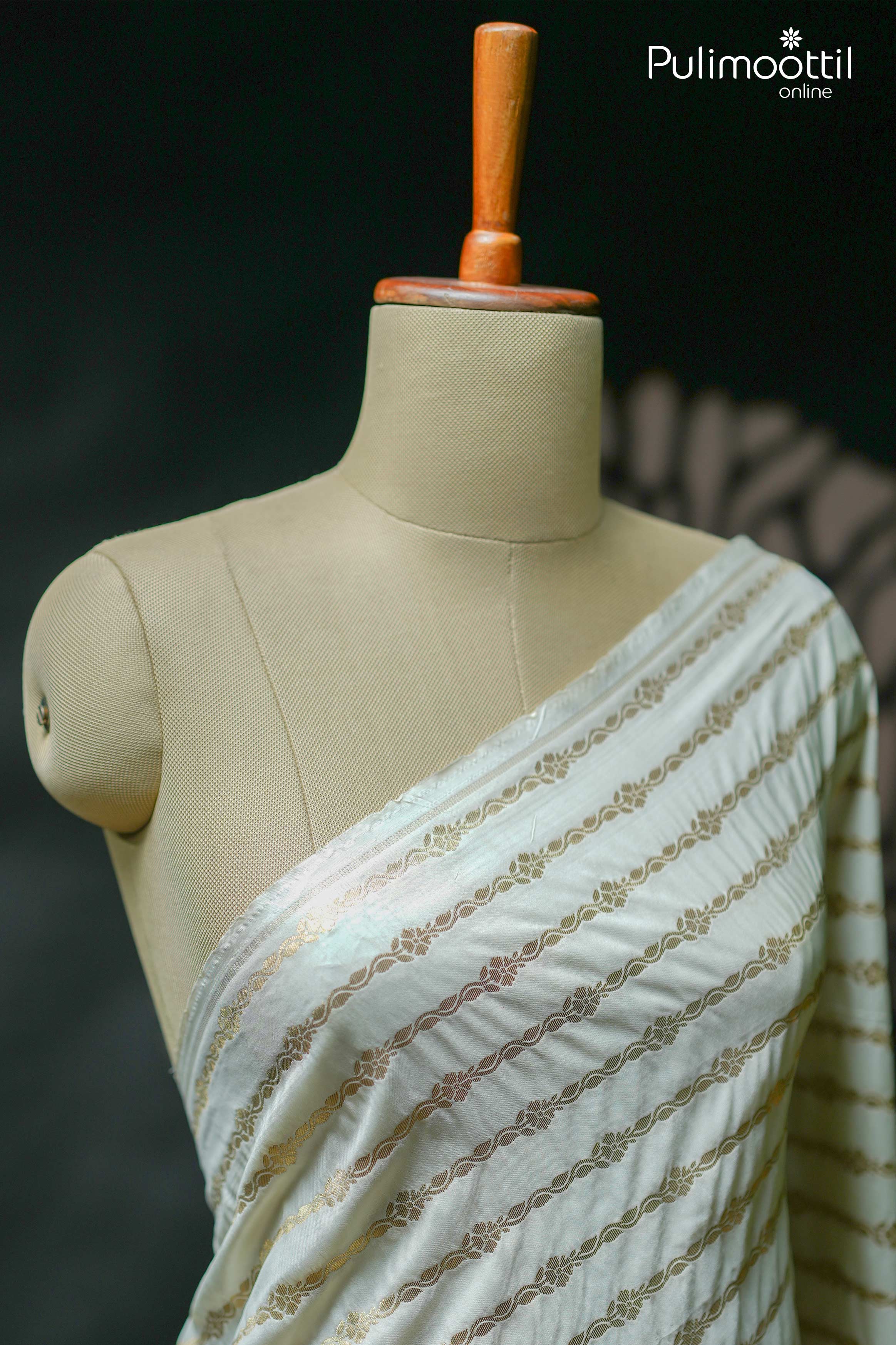 Close-up of off-white semi-silk saree with golden zari floral stripes and subtle sheen, draped over mannequin shoulder to highlight the contrast border design.