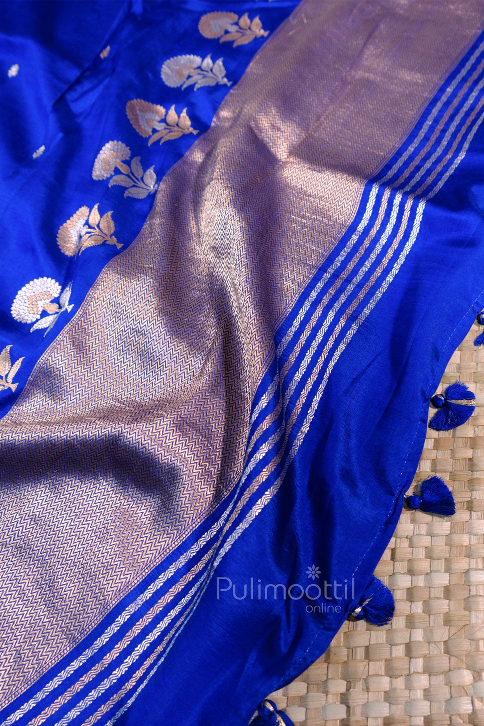 Blue silk saree with golden floral weaving and shimmering zari border featuring zigzag patterns and blue tassels.