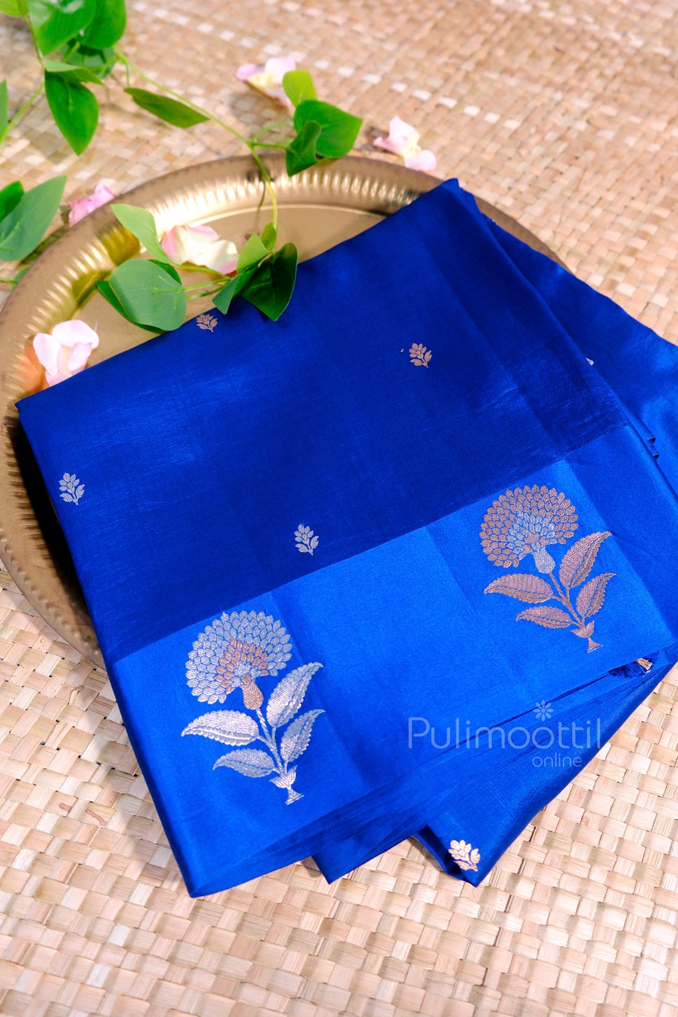 Royal blue silk saree with golden floral motifs and small leaf buttas, displayed on a golden plate with floral decor.