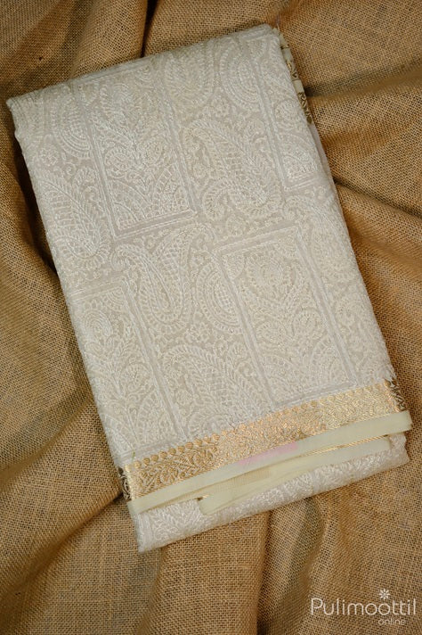 Folded cream net cotta saree with detailed mango floral thread embroidery and golden zari creeper design border, placed on a jute fabric backdrop.