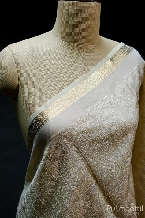 Draped view of cream net cotta saree on mannequin highlighting golden zari creeper border and intricate mango floral thread embroidery.