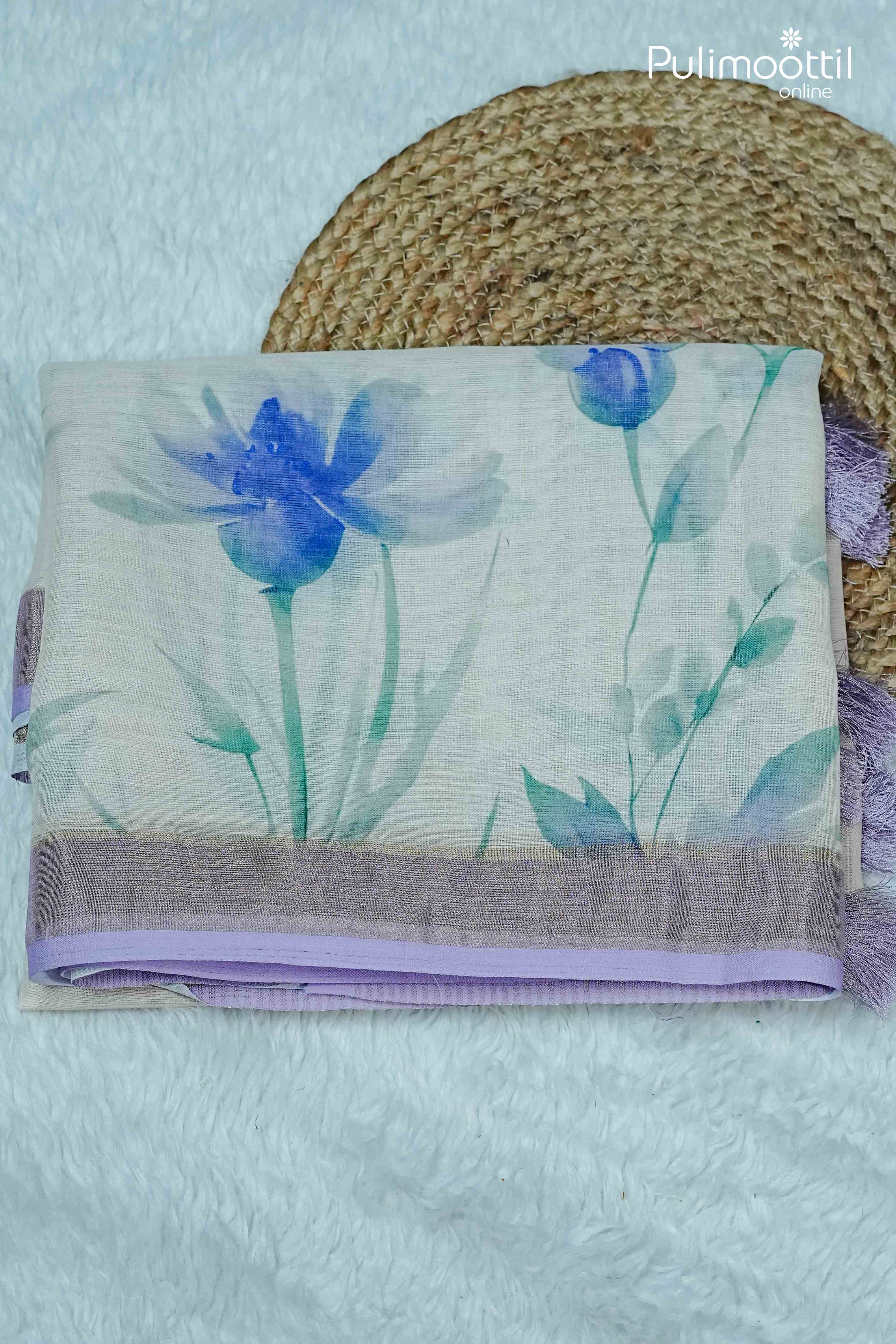 Folded cream semi linen saree featuring blue and green floral prints with a contrast lavender border, displayed on a textured mat – Pulimoottil Online