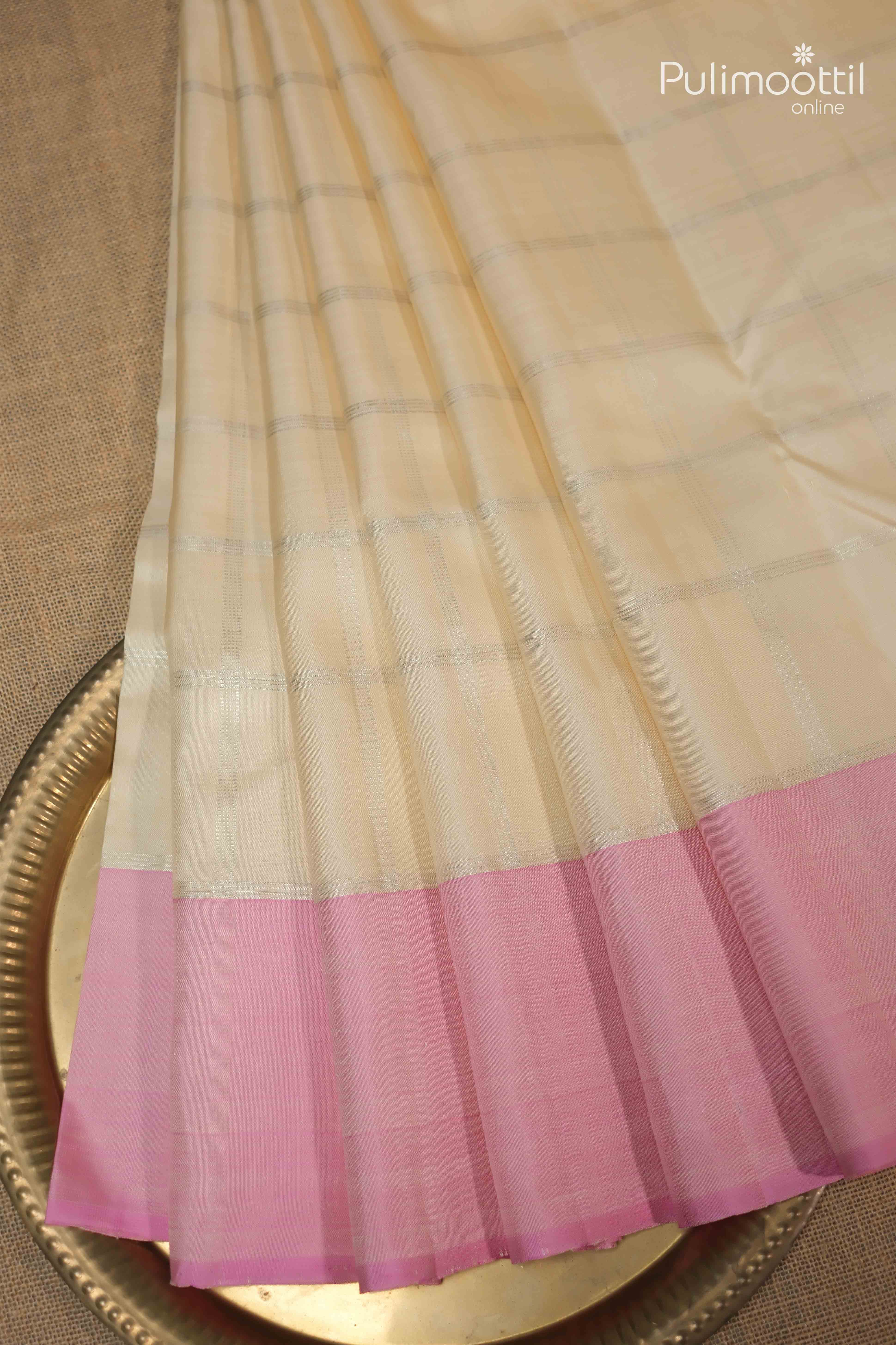 Cream colour pure Kanchipuram silk saree with silver checked weave and soft pink border, neatly pleated and displayed on a brass plate.