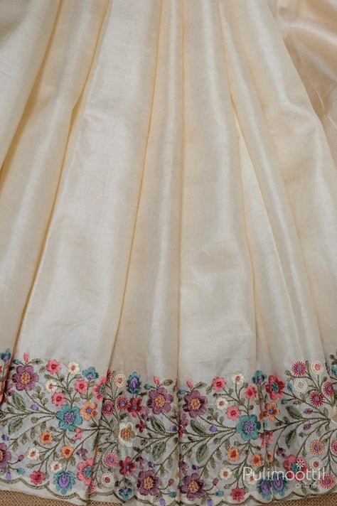 Cream-colored soft silk saree with multicolor floral embroidery on the border featuring intricate threadwork in pink, purple, blue, and green hues.