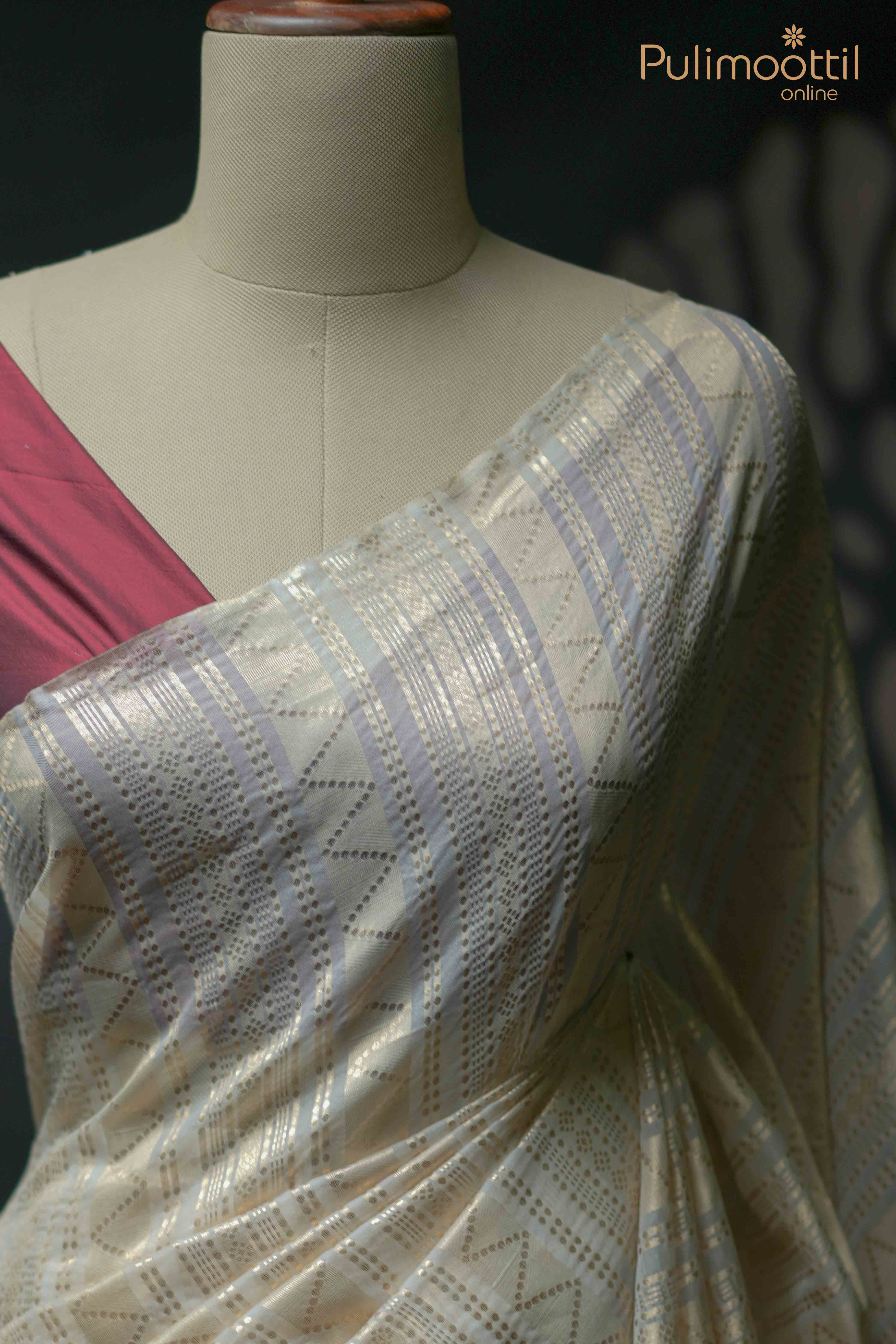 Cream organza saree with golden zari geometric pattern draped on mannequin, paired with contrasting maroon blouse fabric.