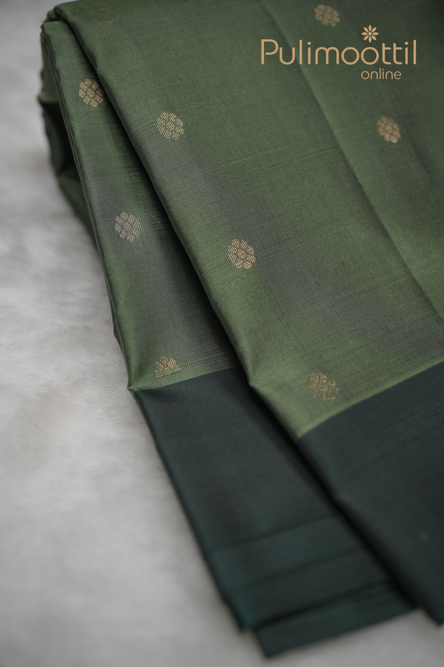 Crocodile green Kanchipuram silk saree with all-over small gold zari buttas and a dark contrast border, neatly folded to showcase its rich texture and traditional weave.