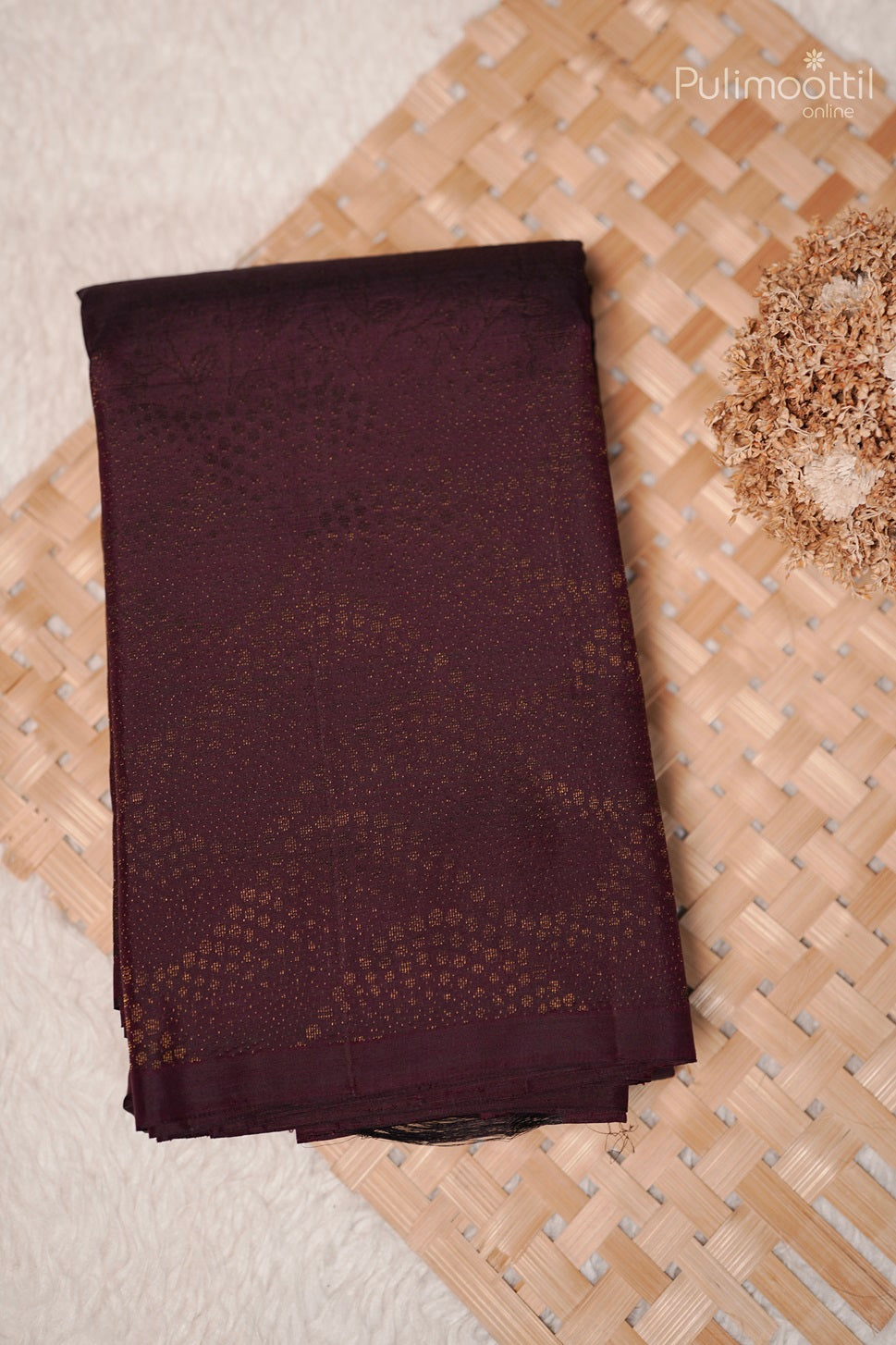 Folded dark purple Kanchipuram designer saree placed on a woven mat with dried flowers for decoration.