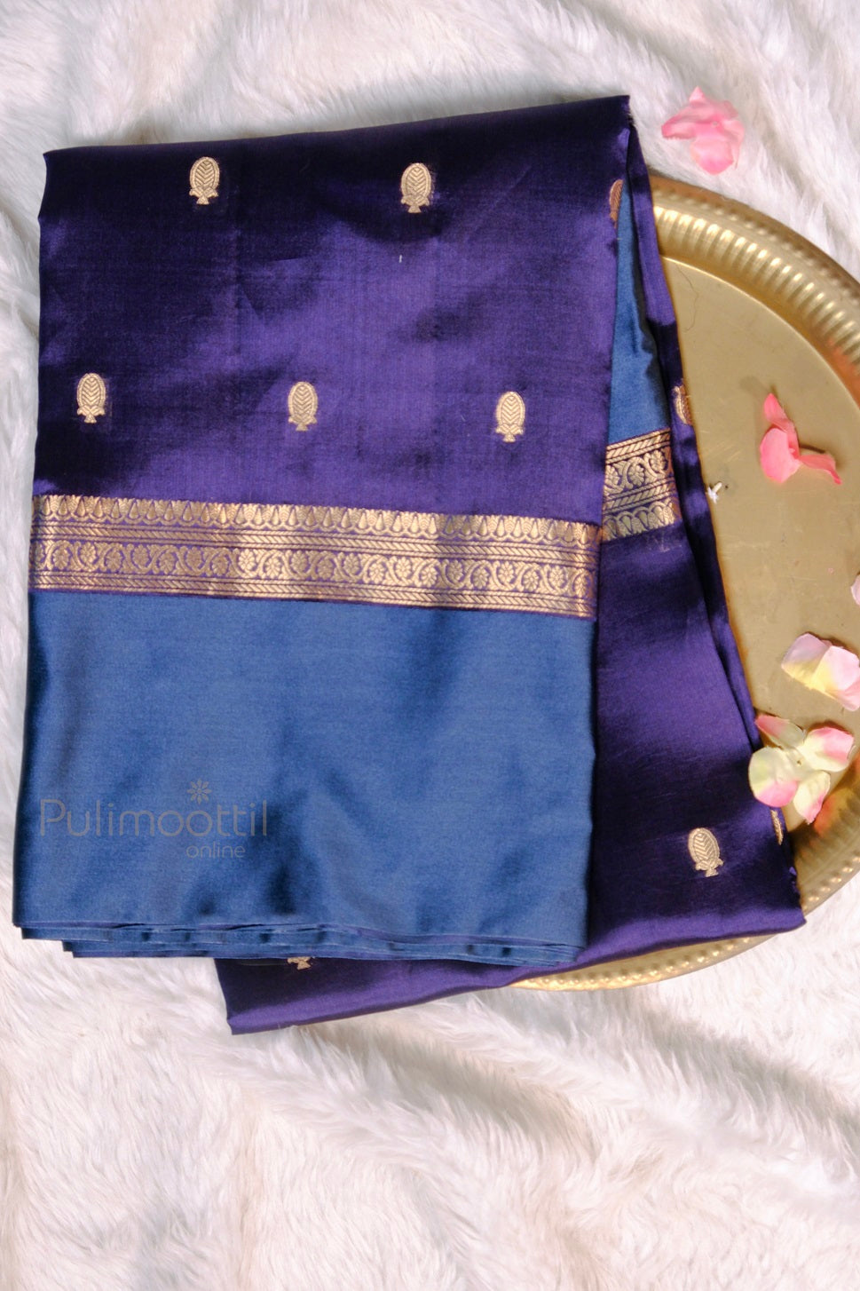 Folded mango silk saree in deep purple and blue shades with elegant gold zari butta and border, laid on a white fur background with flower petals.