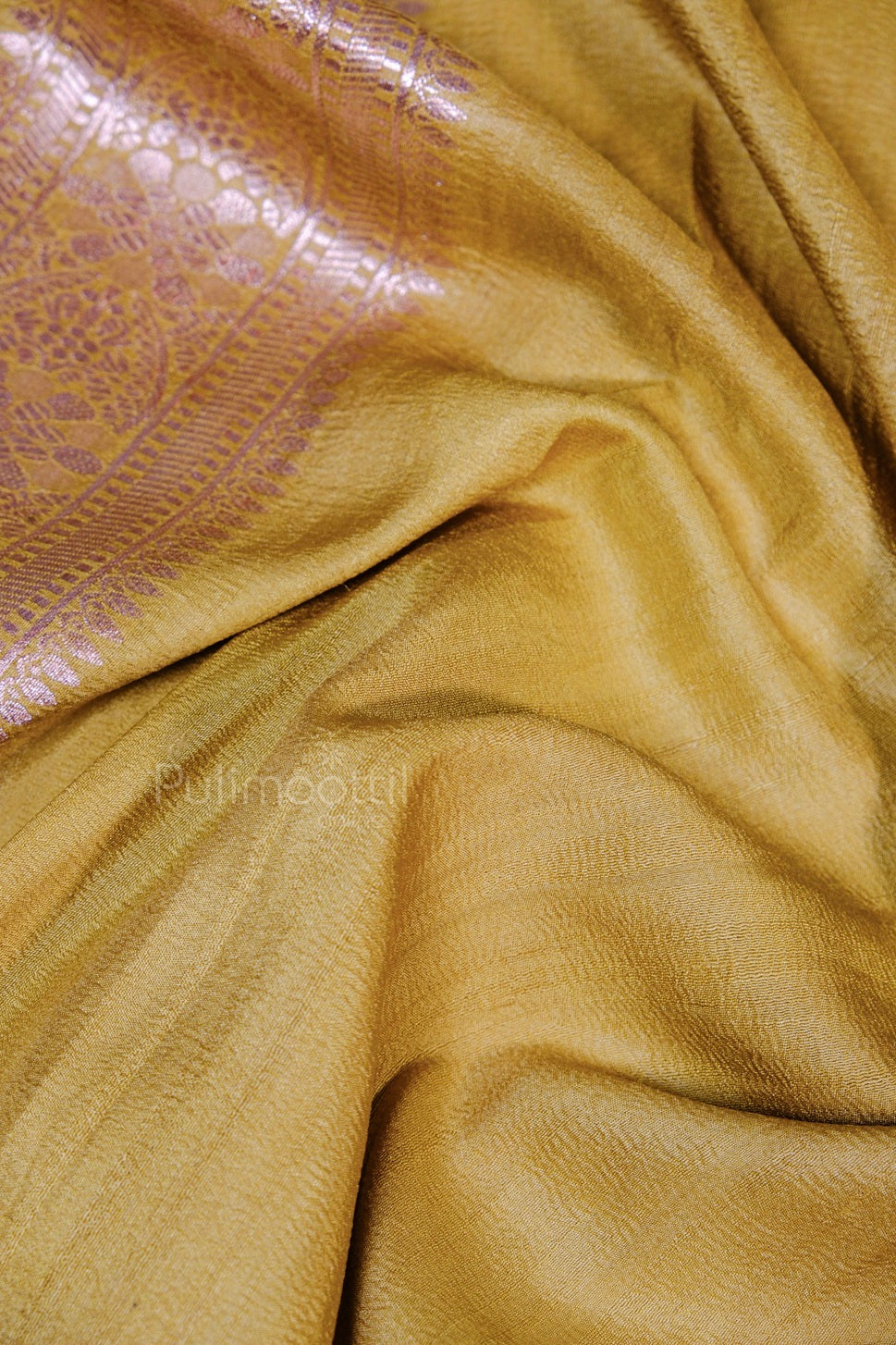Folded golden silk saree with subtle floral and paisley jacquard border in a shimmering mauve-gold tone.