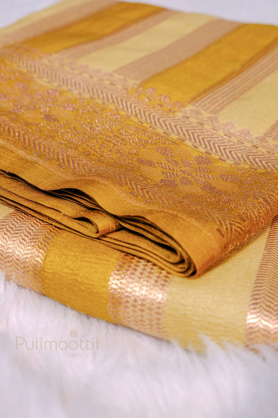 Folded golden silk saree laid on white tile background; paisley and chevron jacquard in mauve‑gold running along stripes.