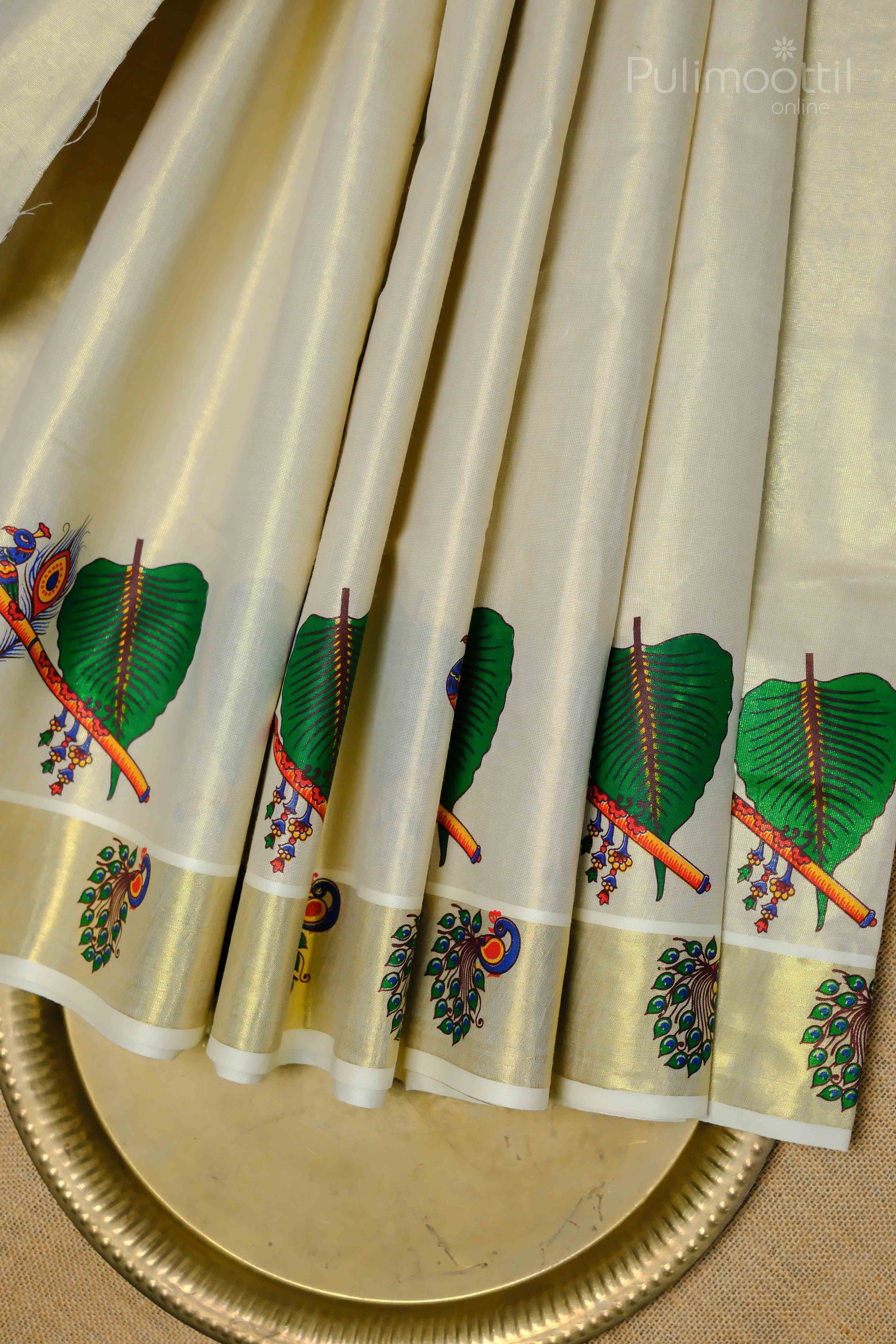 Cream‑gold Kerala tissue saree draped to show its shimmering golden kasavu border and hand‑painted green peacock mural motif along lower edge and pallu.