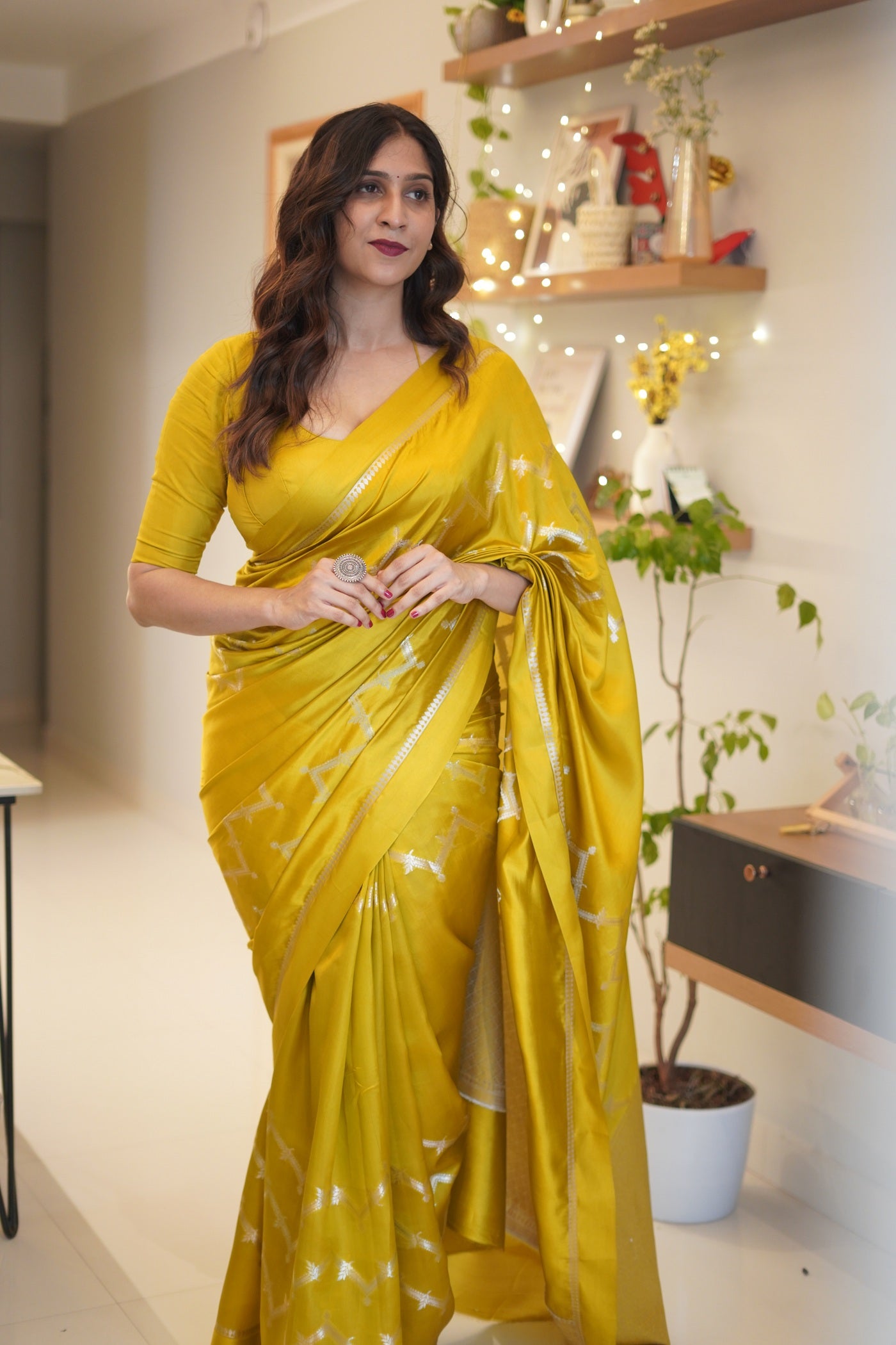 Woman in a mustard yellow silk saree standing indoors with decorative lights and plants in the background.