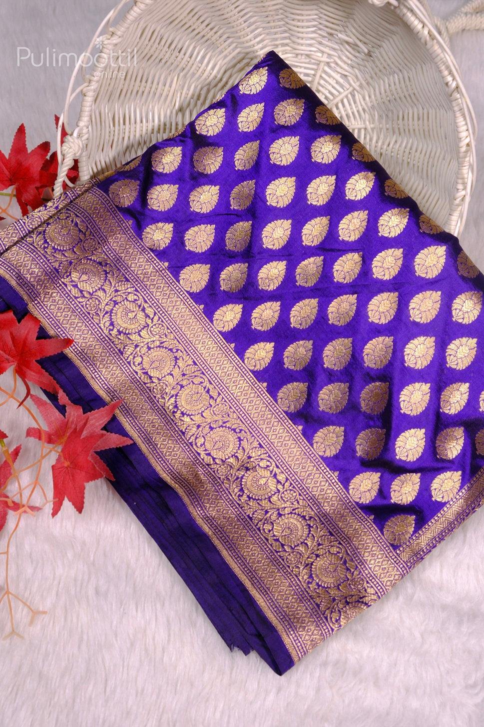 Folded royal purple Banarasi silk saree with gold buti motifs and ornate zari floral border displayed in a white wicker basket.