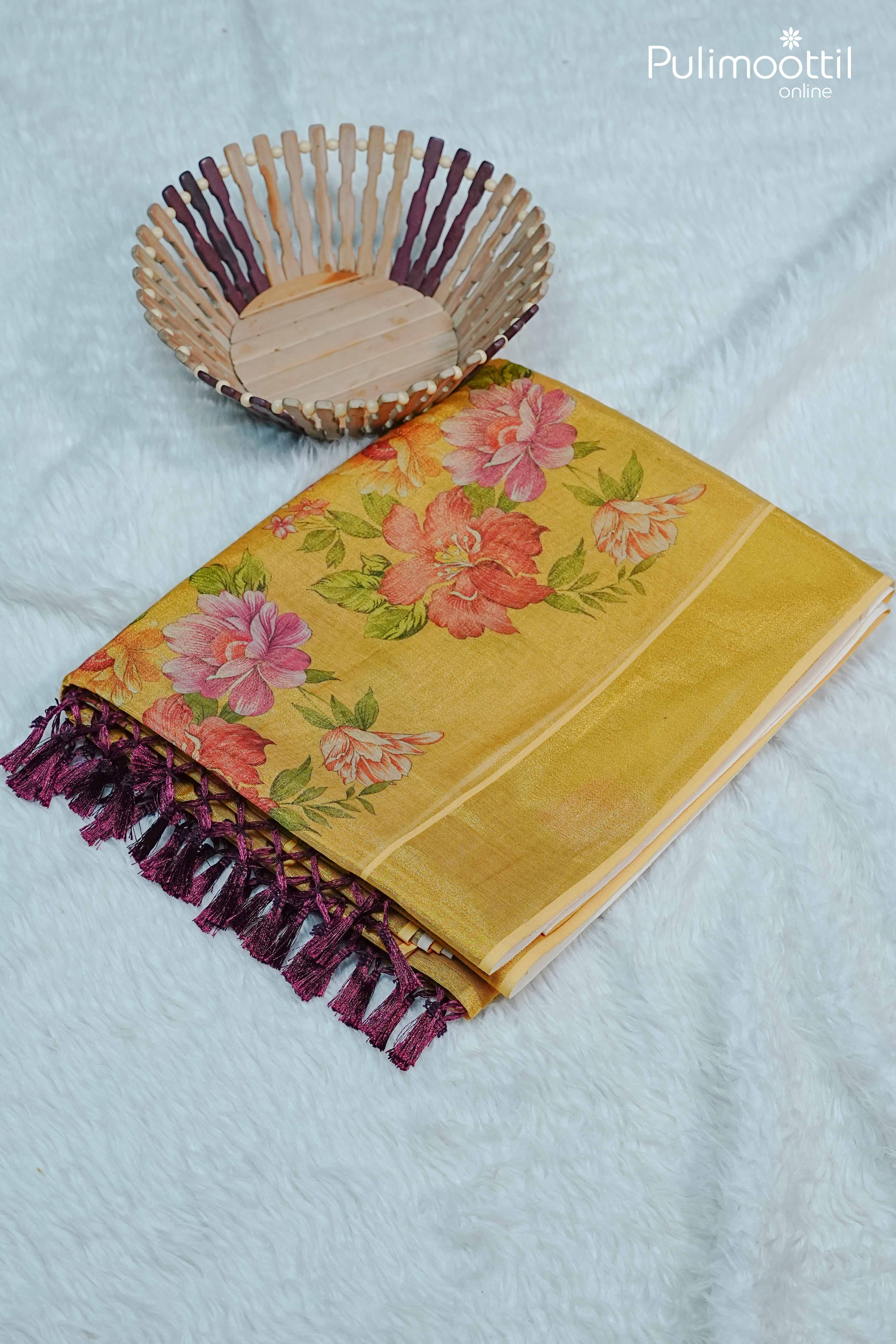 Folded golden yellow Kerala tissue saree with multicolour floral print and maroon tassel edging, styled on white fur background with wicker basket.