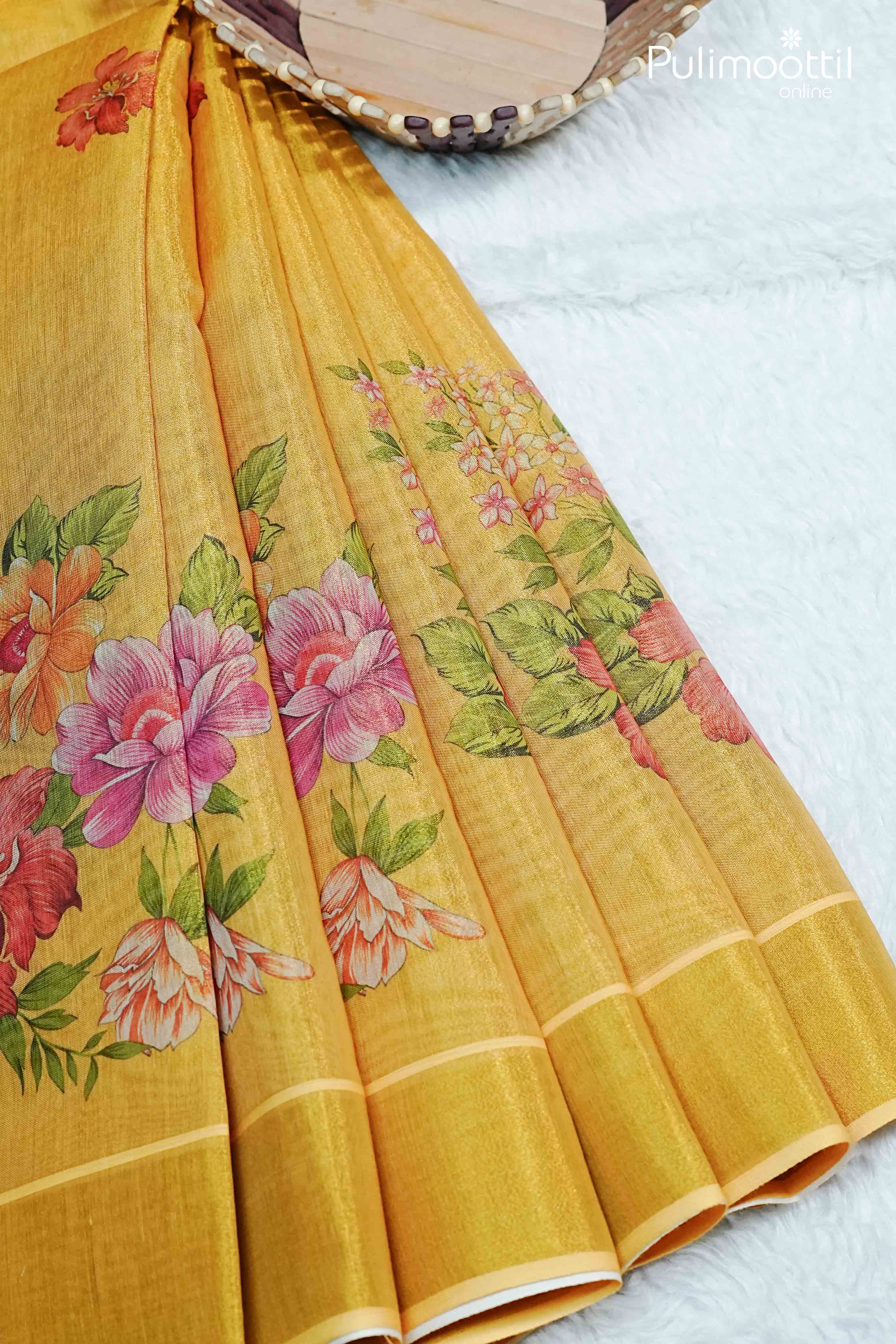 Pleated golden yellow Kerala tissue saree featuring multicolour floral designs along the border and pallu, laid on white textured background.