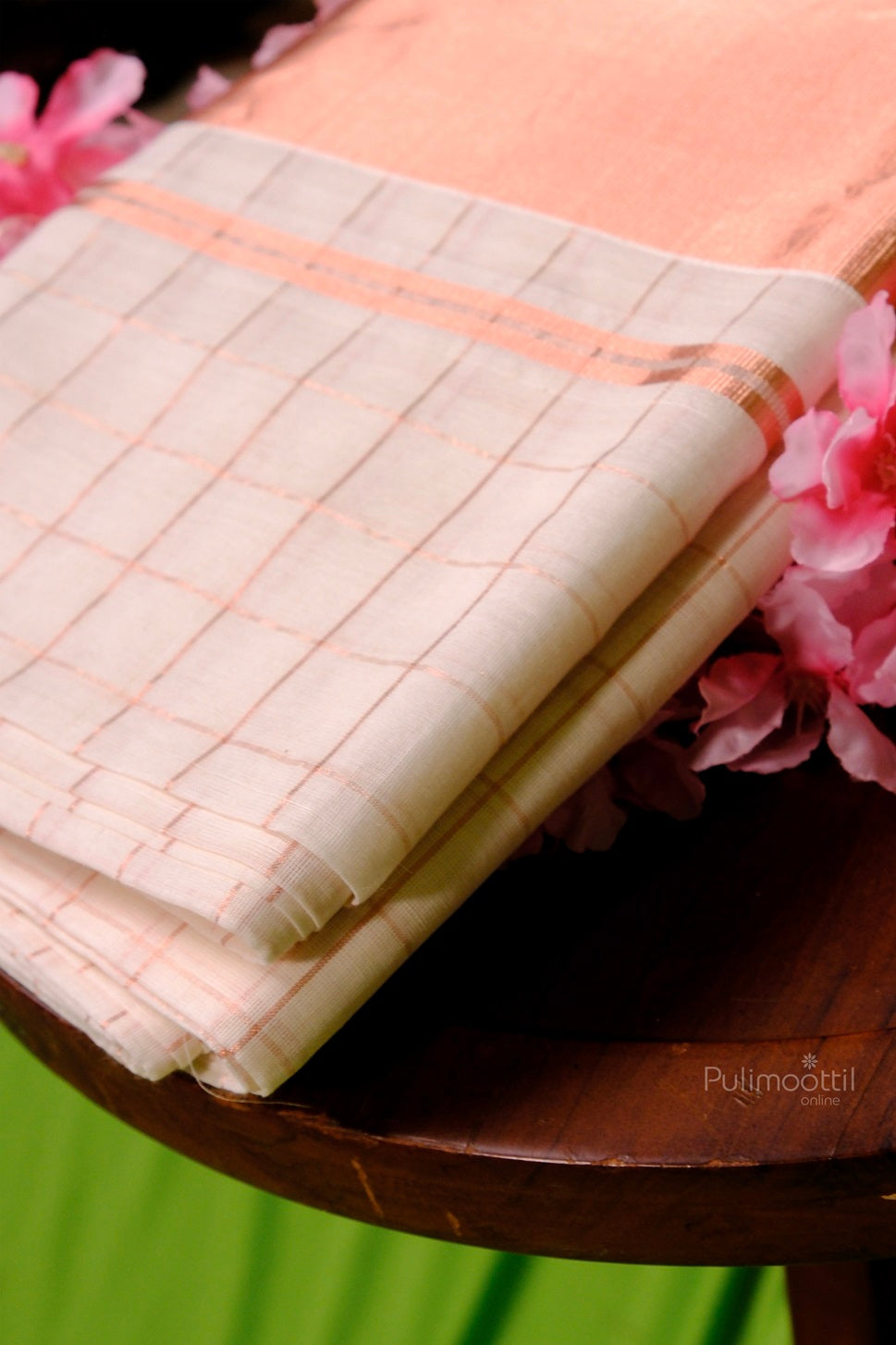 Folded Kerala Kasavu saree with subtle copper checkered design and shimmering border, placed on a round wooden surface