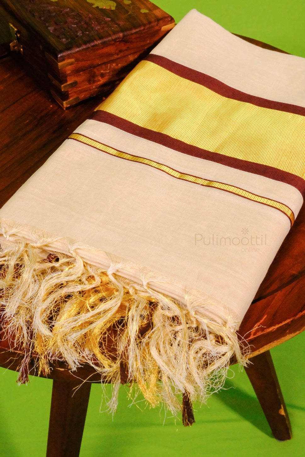 Folded cream-colored Kerala kasavu mundu with broad golden and maroon zari border and fringed edge, placed on a wooden stool.