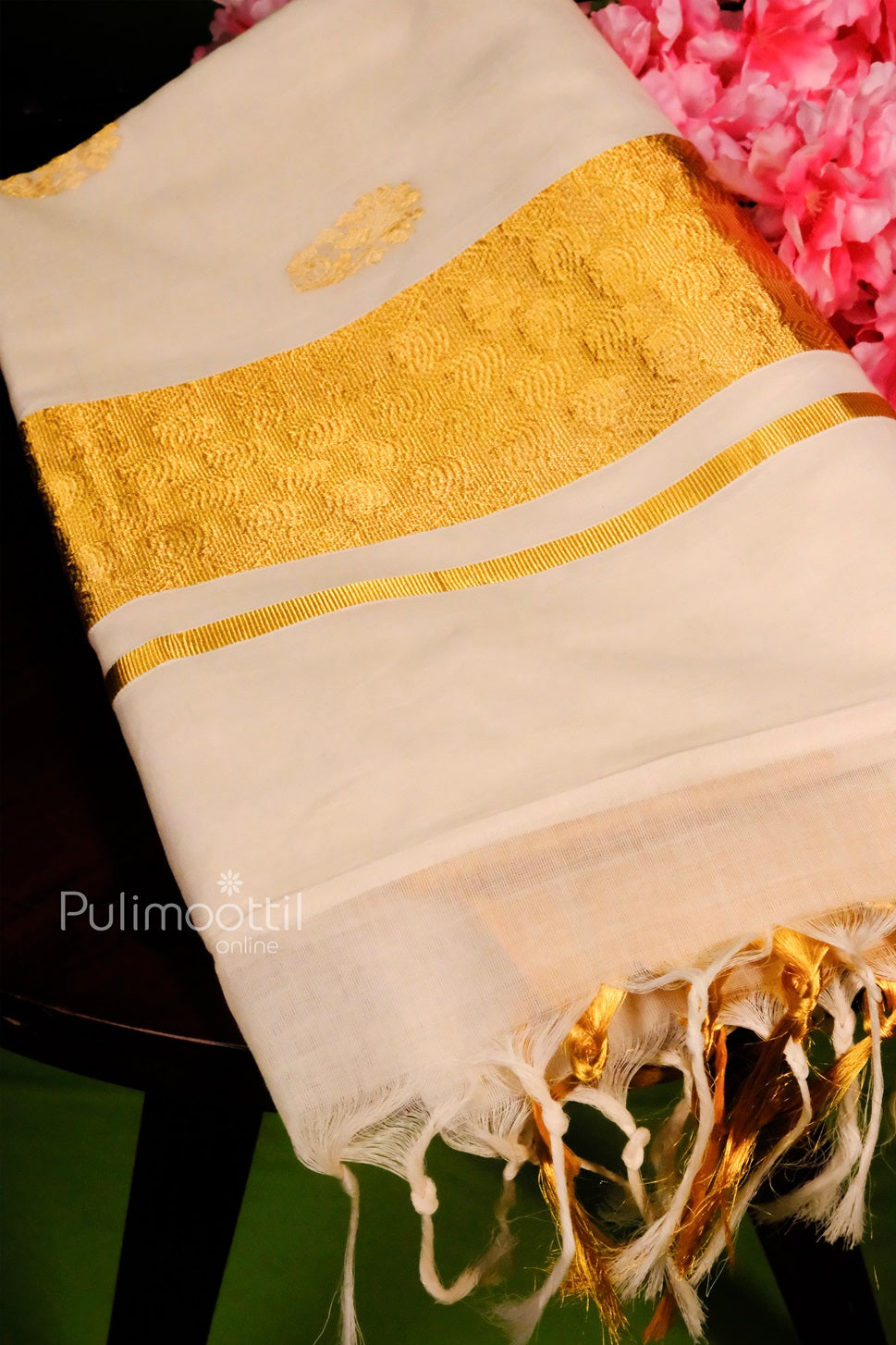 Folded off-white Kerala kasavu saree with golden zari border and tassels, placed beside pink flowers.
