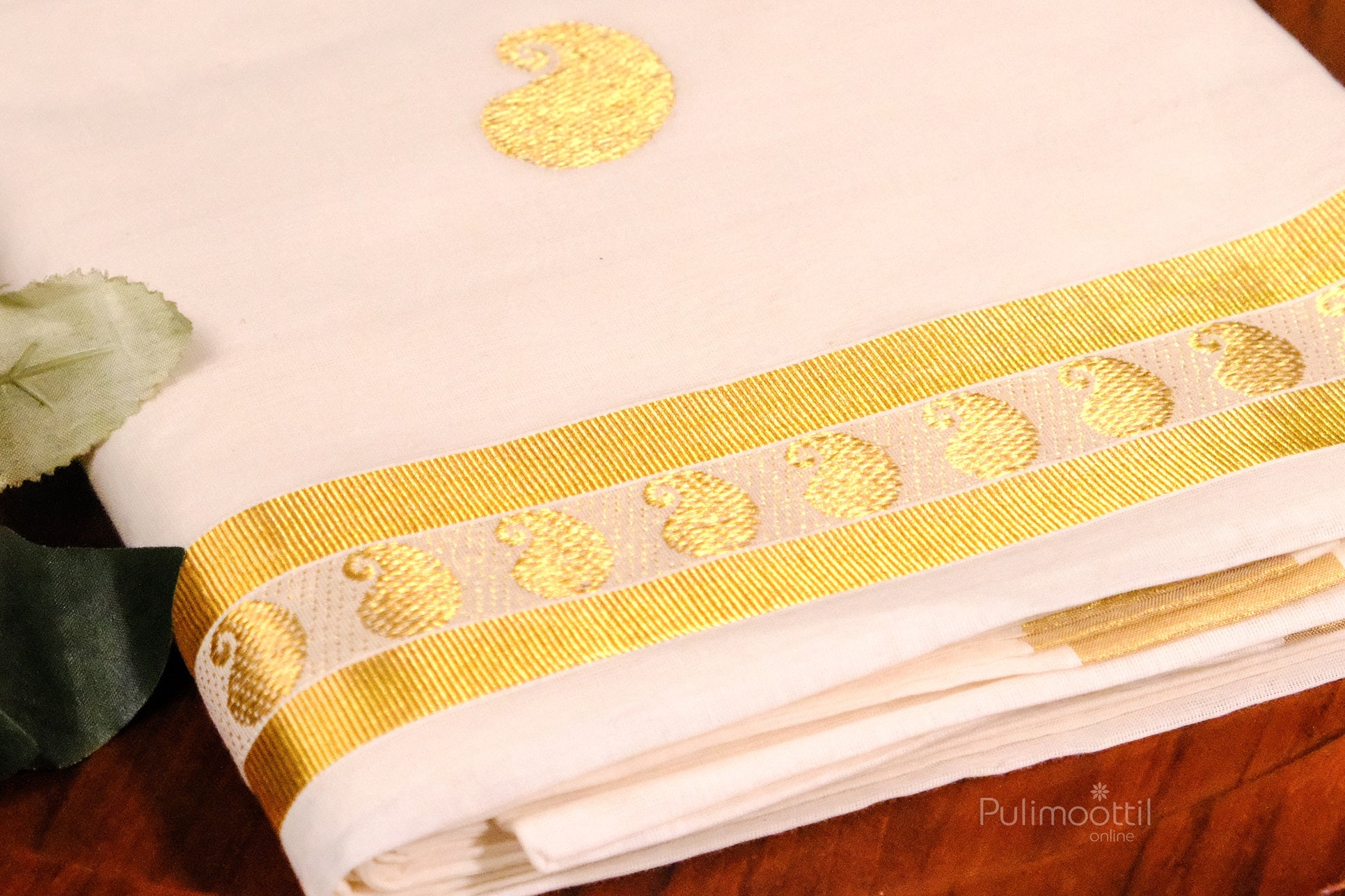 Neatly folded Kerala saree with repeated golden paisley motifs and a detailed gold zari border, adorned with nearby green leaves and flowers.
