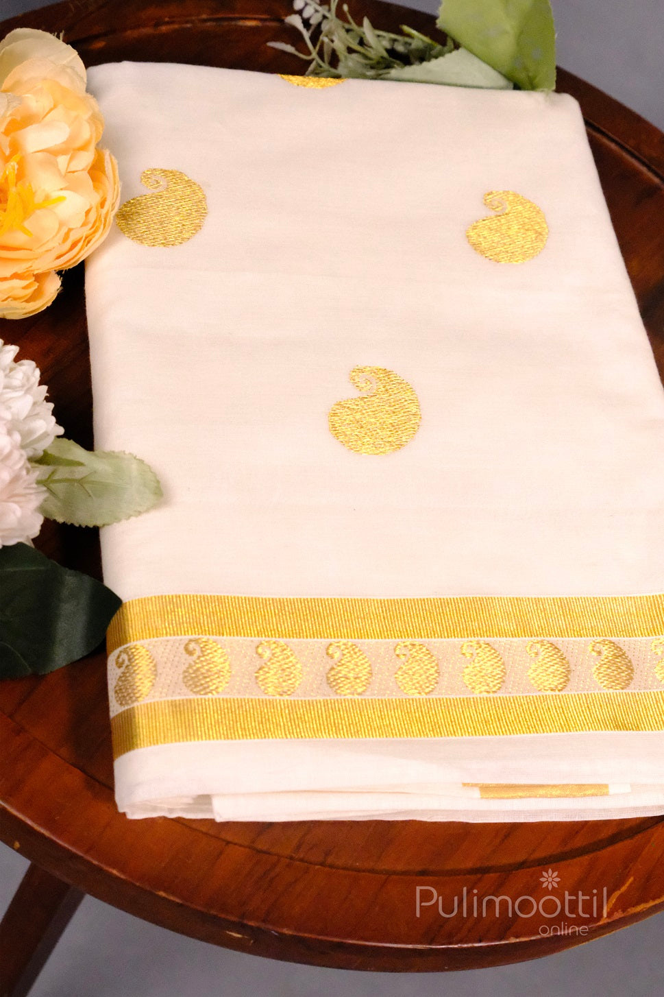Off-white traditional Kerala kasavu saree with golden paisley motifs and a golden zari border, placed on a wooden table with flowers.
