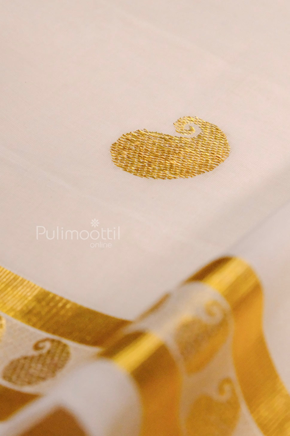 Close-up of a golden paisley motif on an off-white Kerala saree, showing detailed zari embroidery and part of the golden border.