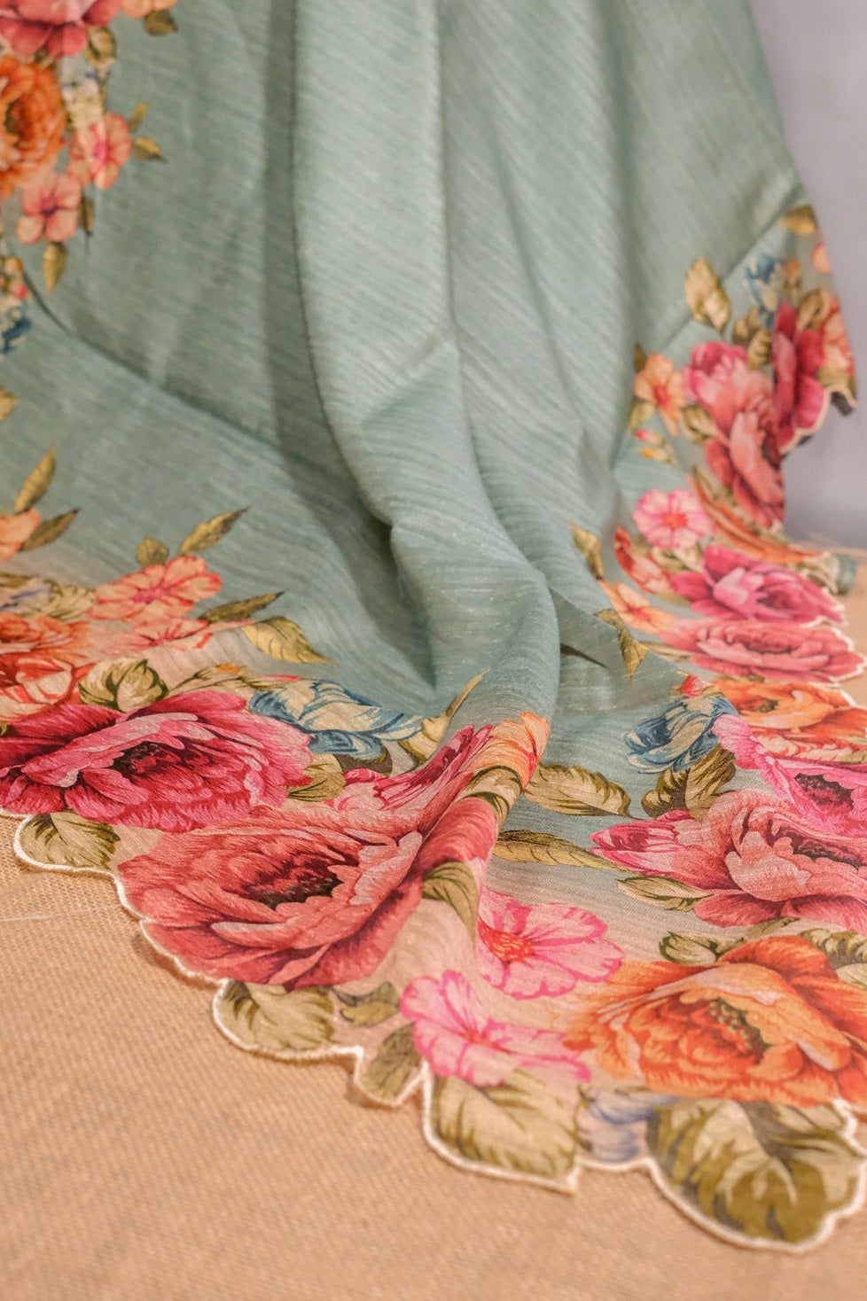 Elegant drape of light teal semi jute saree with multicolored floral border and scalloped edges.
