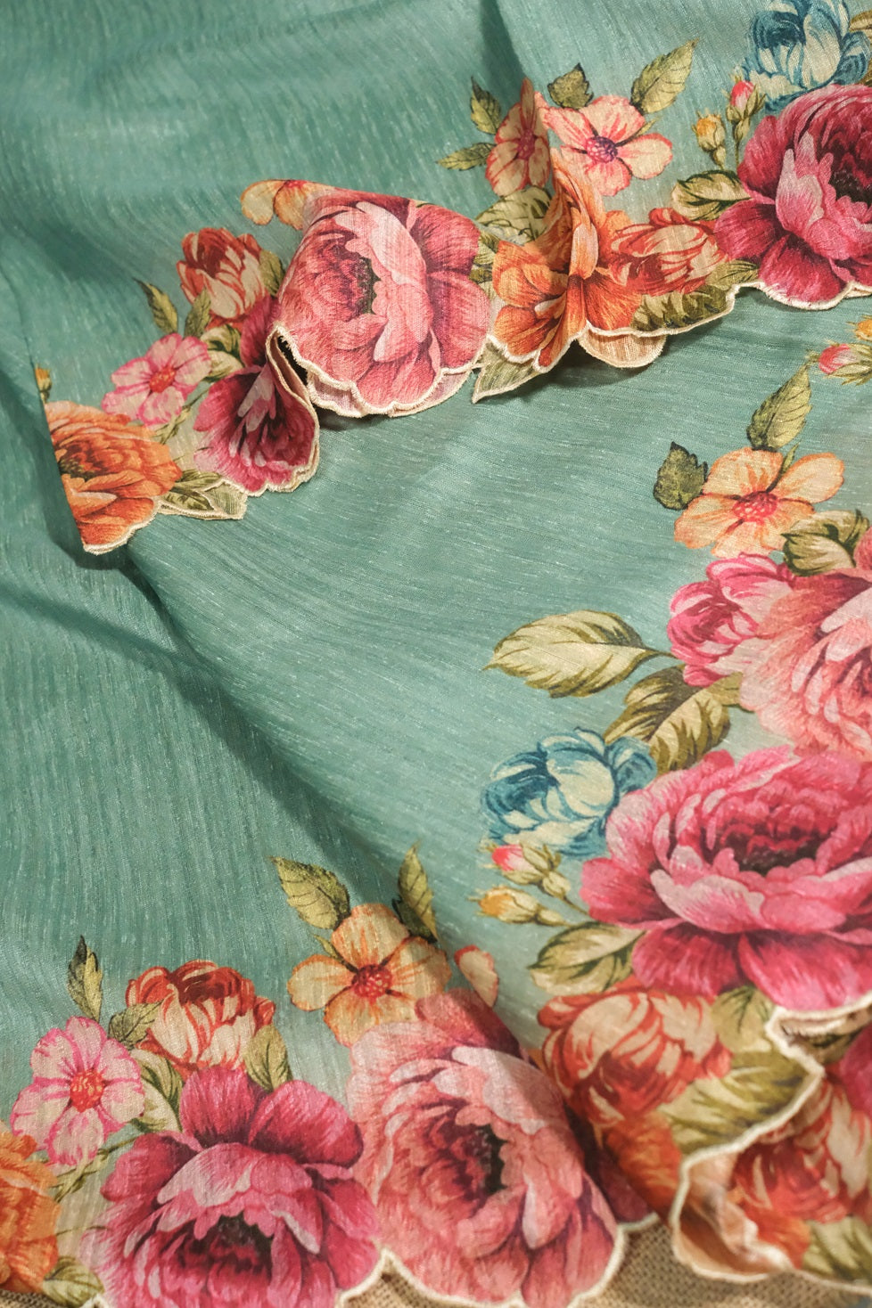 Elegant drape of light teal semi jute saree with multicolored floral border and scalloped edges.