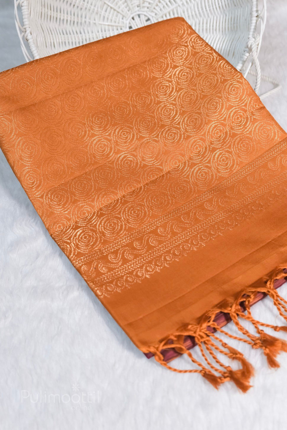 Magenta‑pink borderless Kanchipuram soft silk saree with ikkat pattern and gold‑zari floral motifs, elegantly draped on white faux‑fur in front of a wicker chair