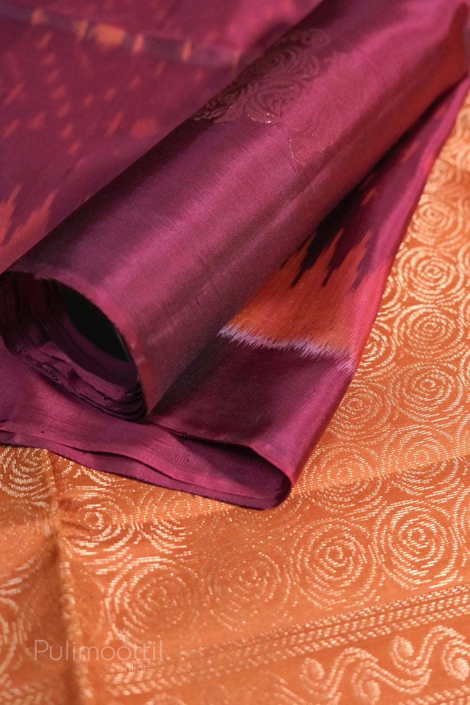 Magenta-pink Kanchipuram soft silk fabric with ikkat weave and gold zari floral motifs, partially rolled on orange-gold brocade background