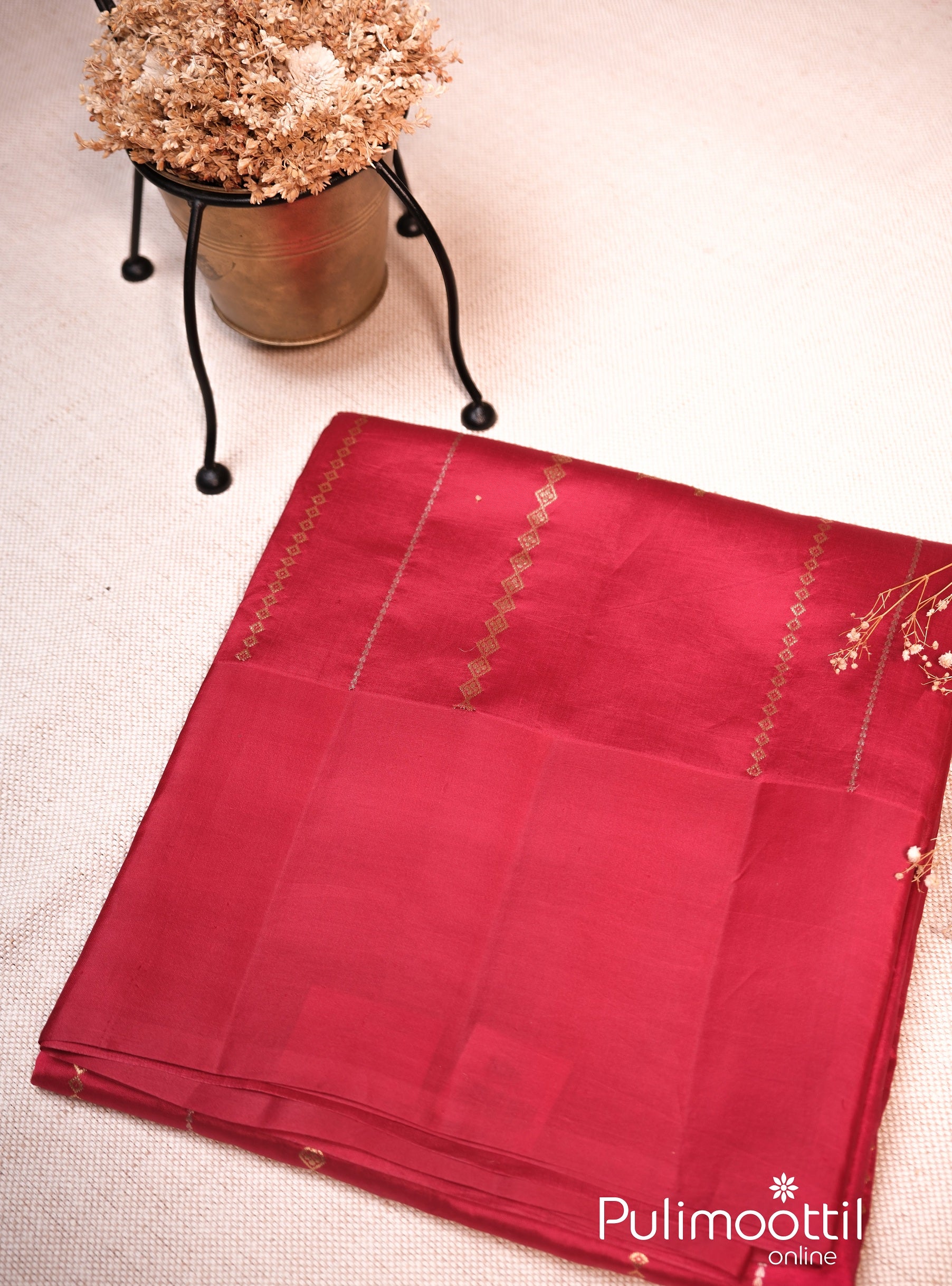 Maroon color mango silk saree neatly folded, featuring subtle golden zari patterns along the border, placed beside a brass pot with dried flowers.