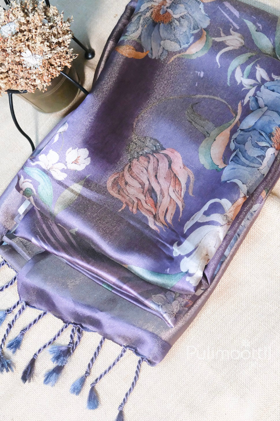 Lavender Color Satin Silk Saree with large floral digital print and tassel pallu, neatly folded and displayed beside a decorative dried flower arrangement.