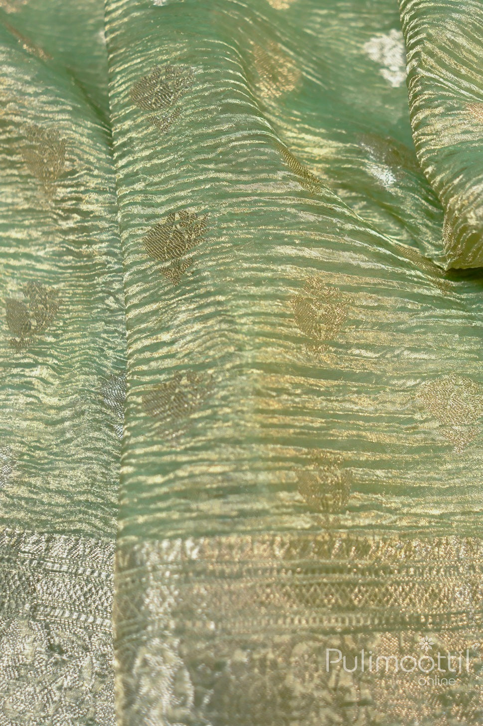 Close-up of mint green Banarasi silk fabric with a subtle crushed texture, featuring delicate gold zari floral buttas and an intricate border, highlighting the luxurious sheen and intricate details of the saree.