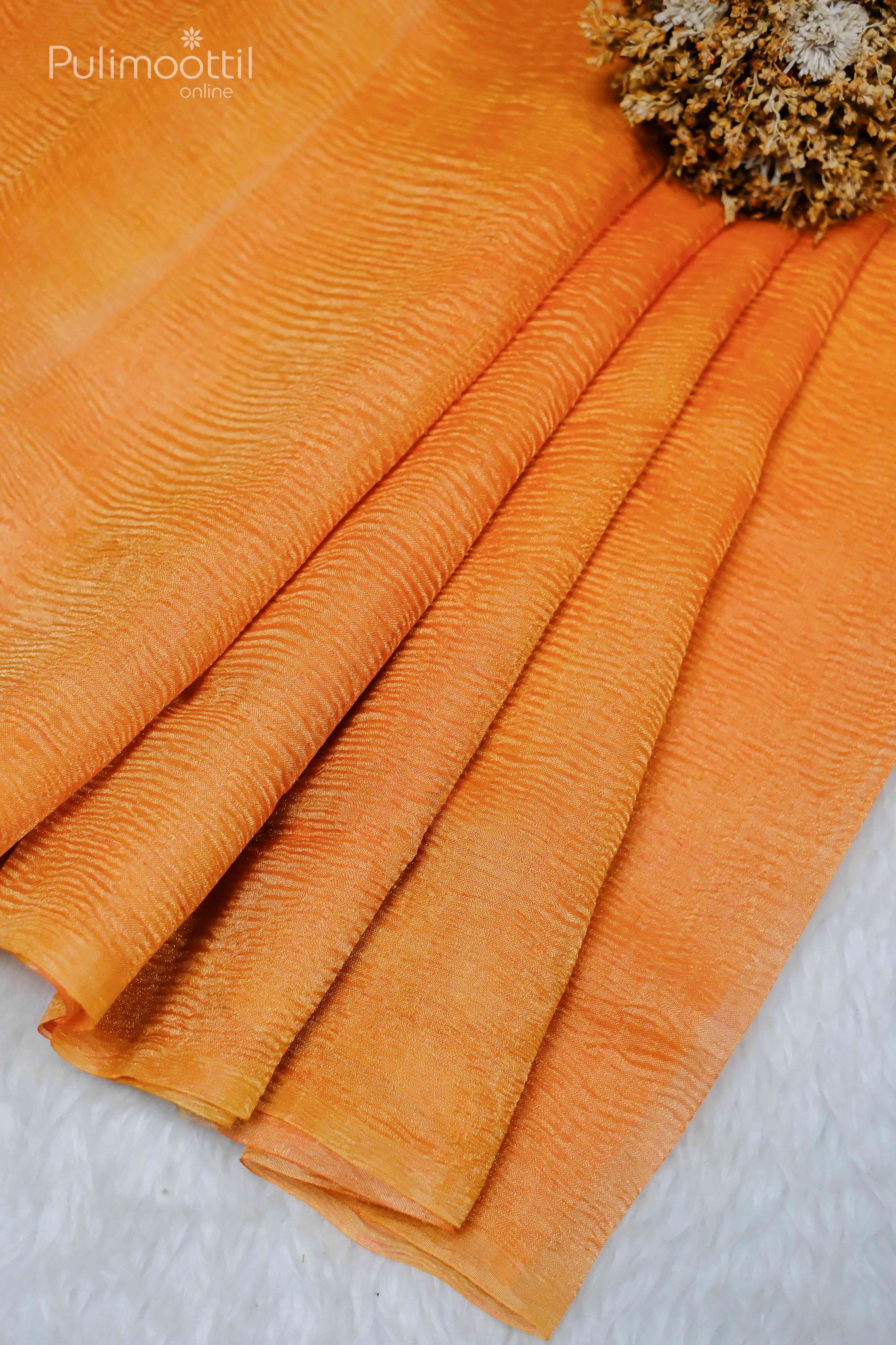 Neon orange crushed tissue saree fabric neatly folded, showcasing its texture, placed on a soft white surface with a potted plant in the background.
