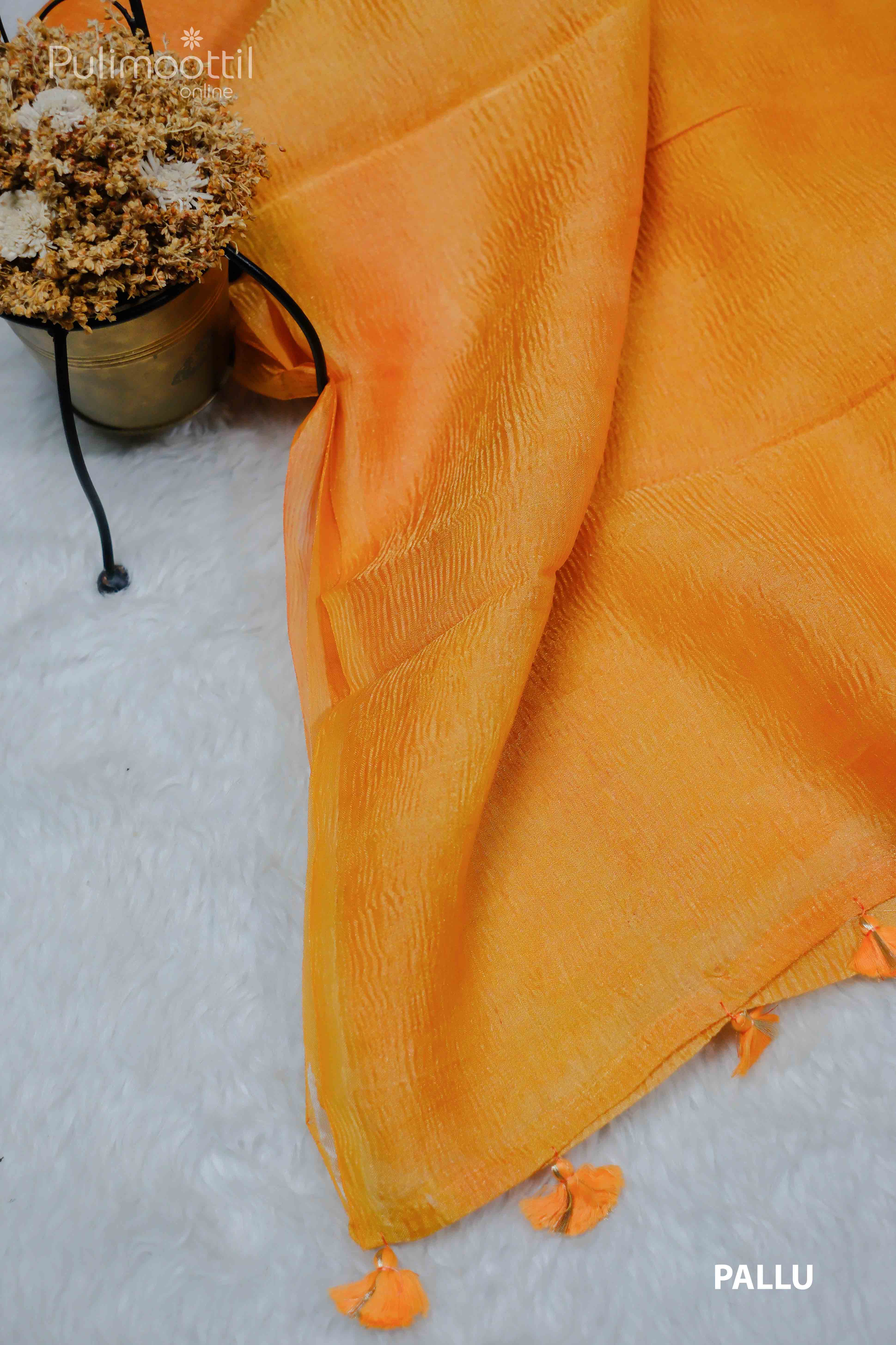 Neon orange crushed tissue saree pallu with tassels, plain without border, placed on a soft white surface with a potted plant in the background.