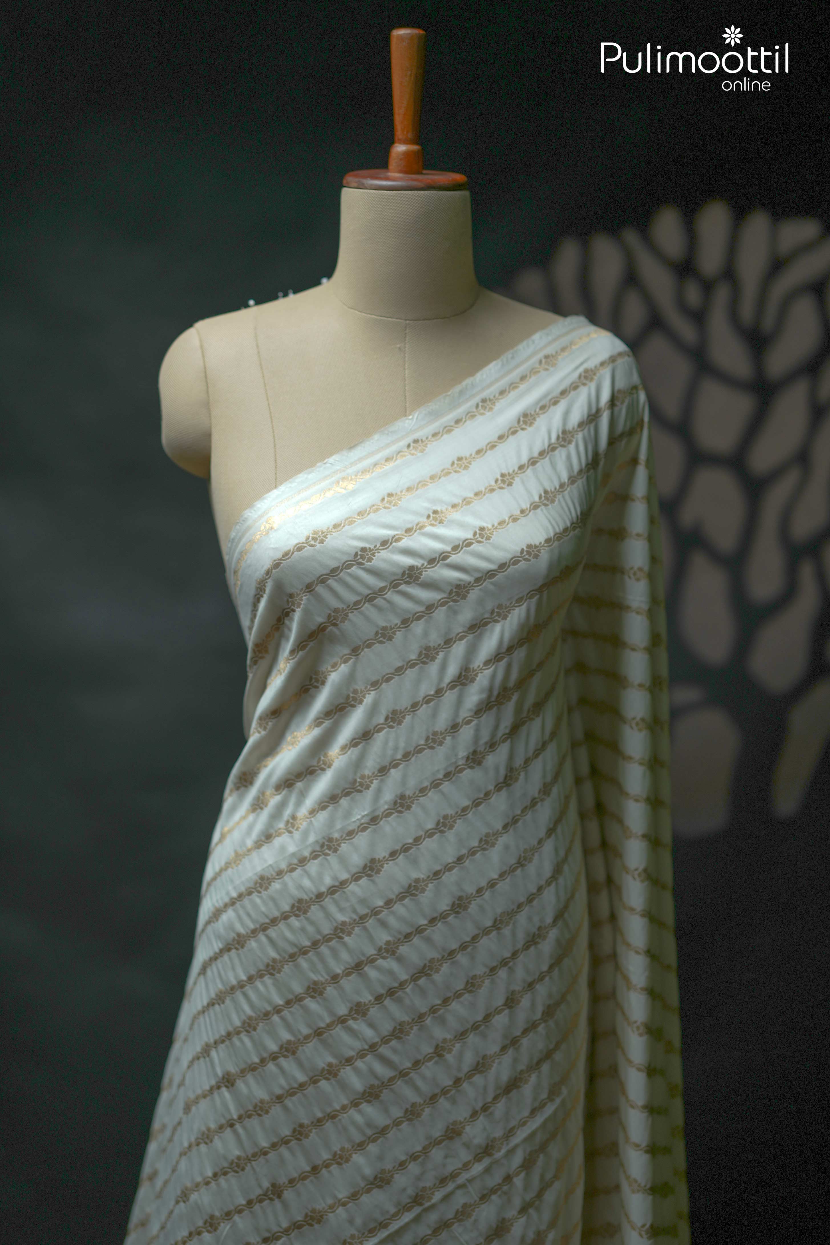 Off-white semi-silk saree draped on mannequin, featuring golden zari floral diagonal stripes and a subtle sheen, shown without blouse or border in view.