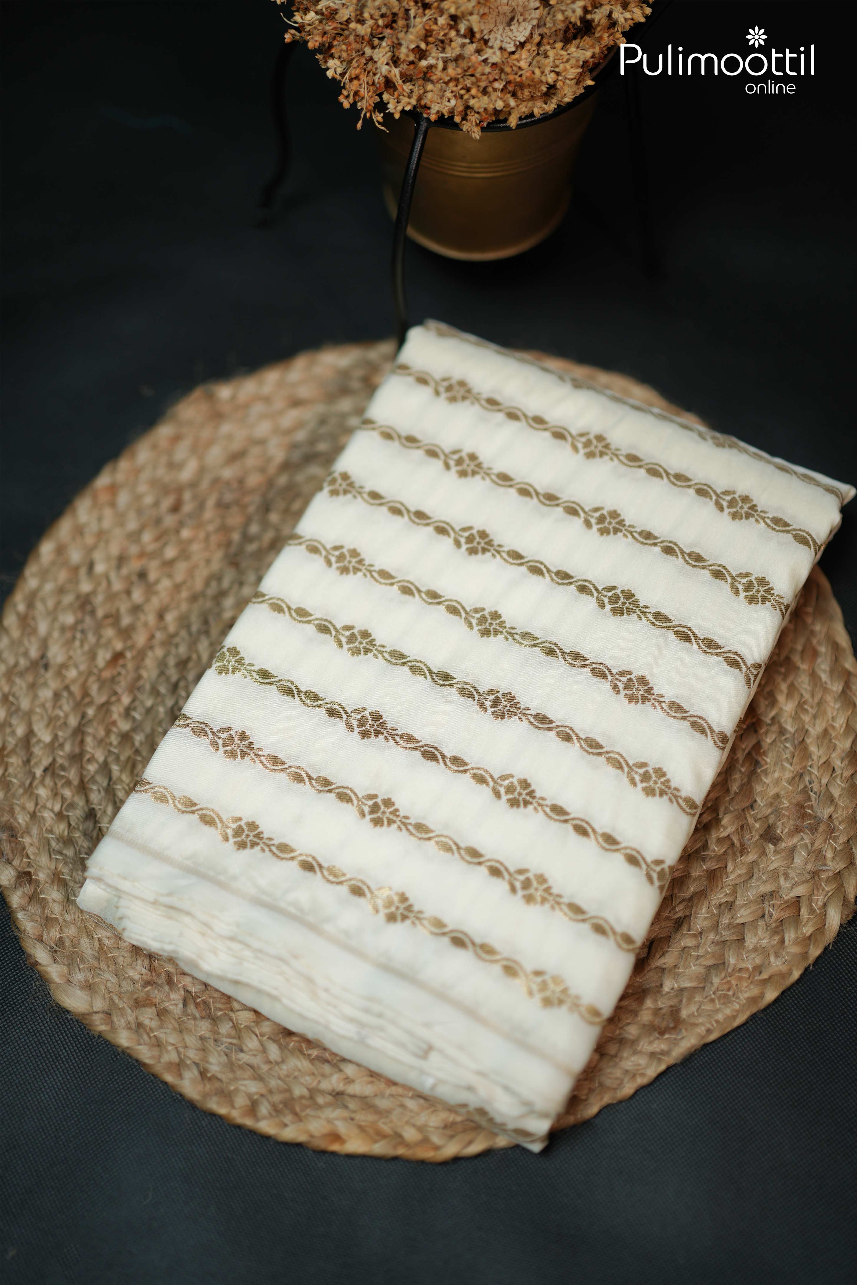 Folded off-white semi-silk saree with golden zari floral stripe pattern, placed on a woven mat with dried flower decor.