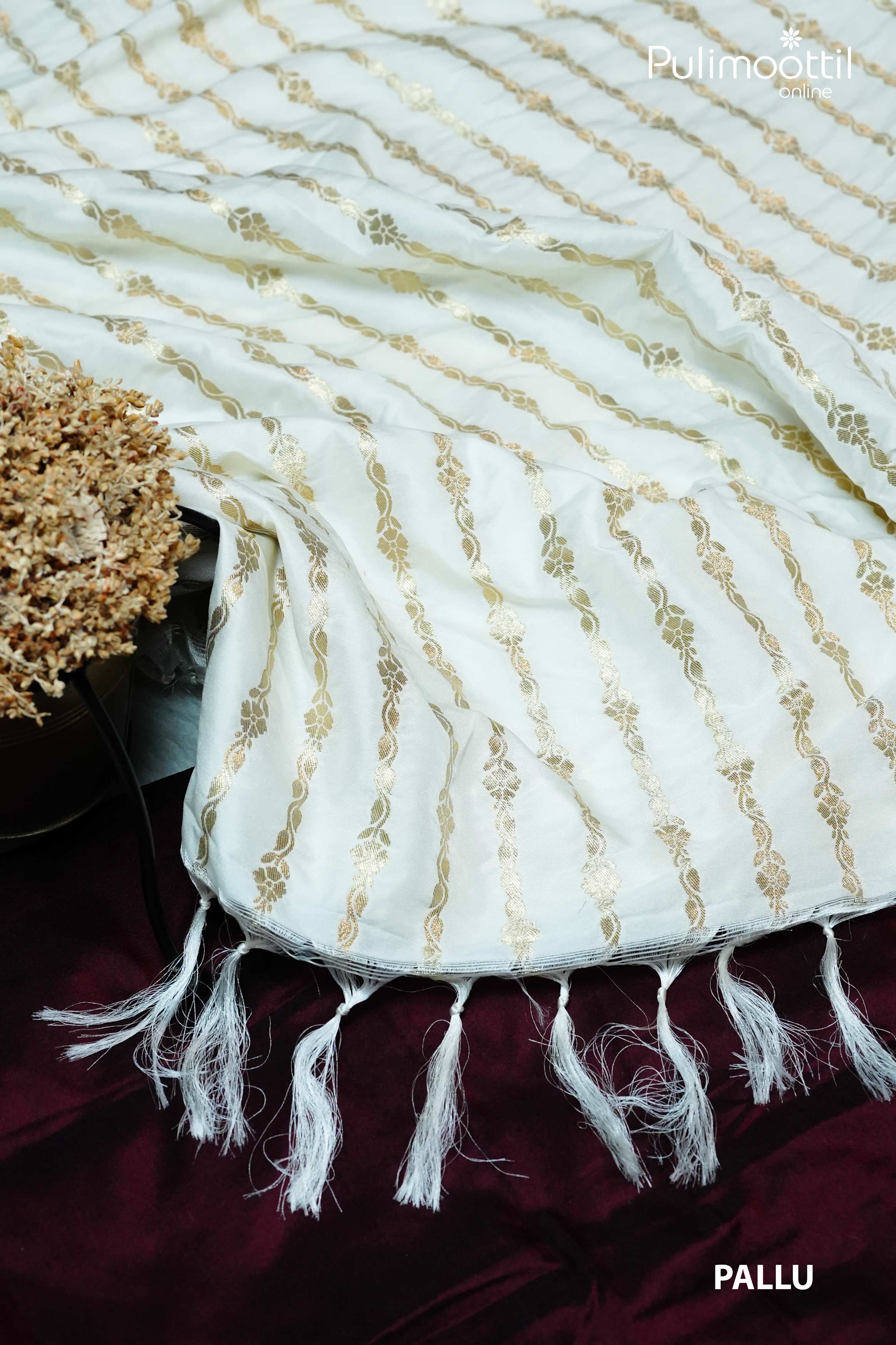 Off-white semi-silk saree pallu with golden zari floral stripes and white tassel edging, displayed on a maroon velvet surface with dried floral decor.