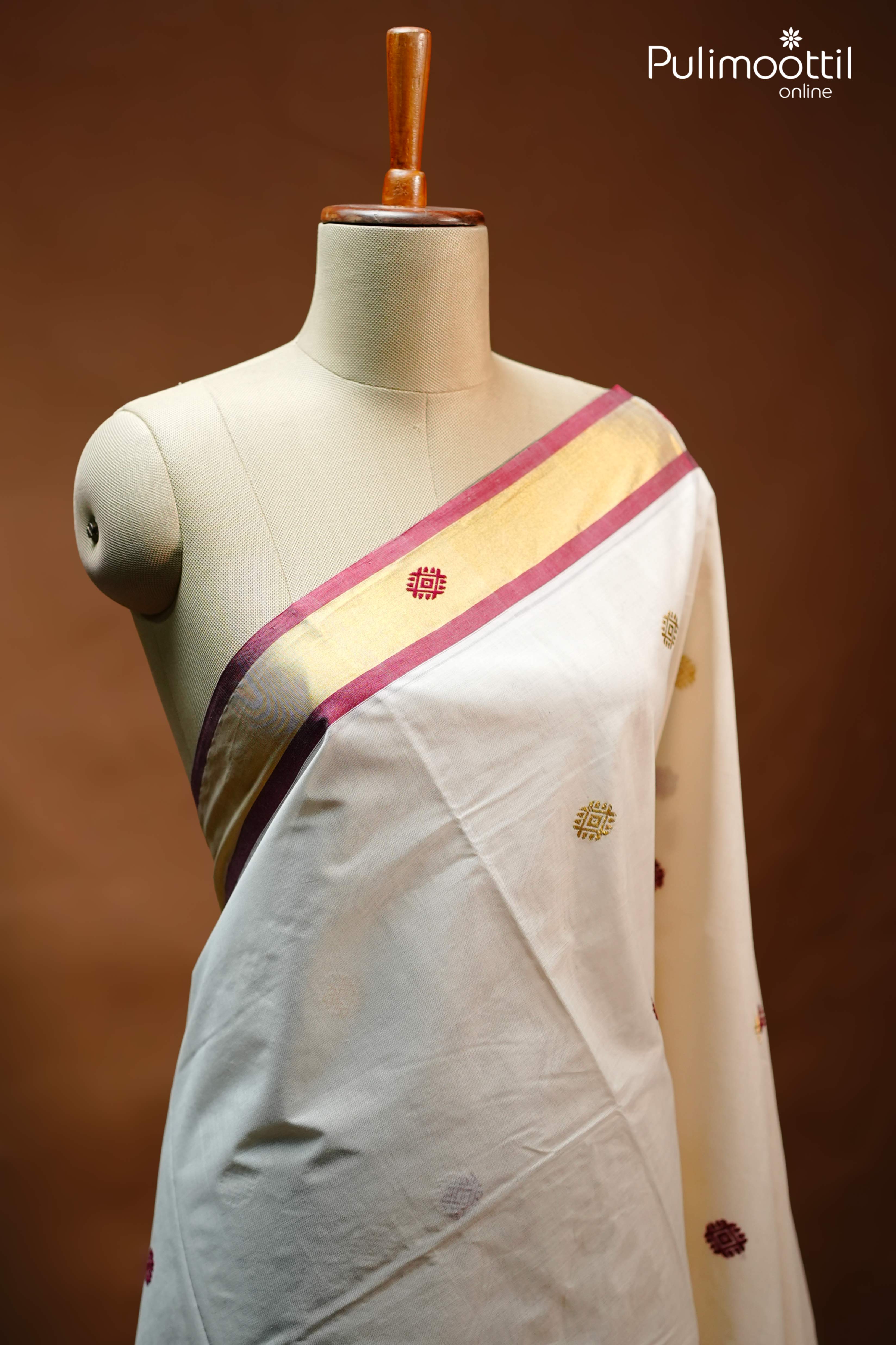 Folded Off White Kerala Set Saree with mixed zari motifs, golden tissue border with maroon edging, and tasselled pallu, placed beside a green tropical leaf.