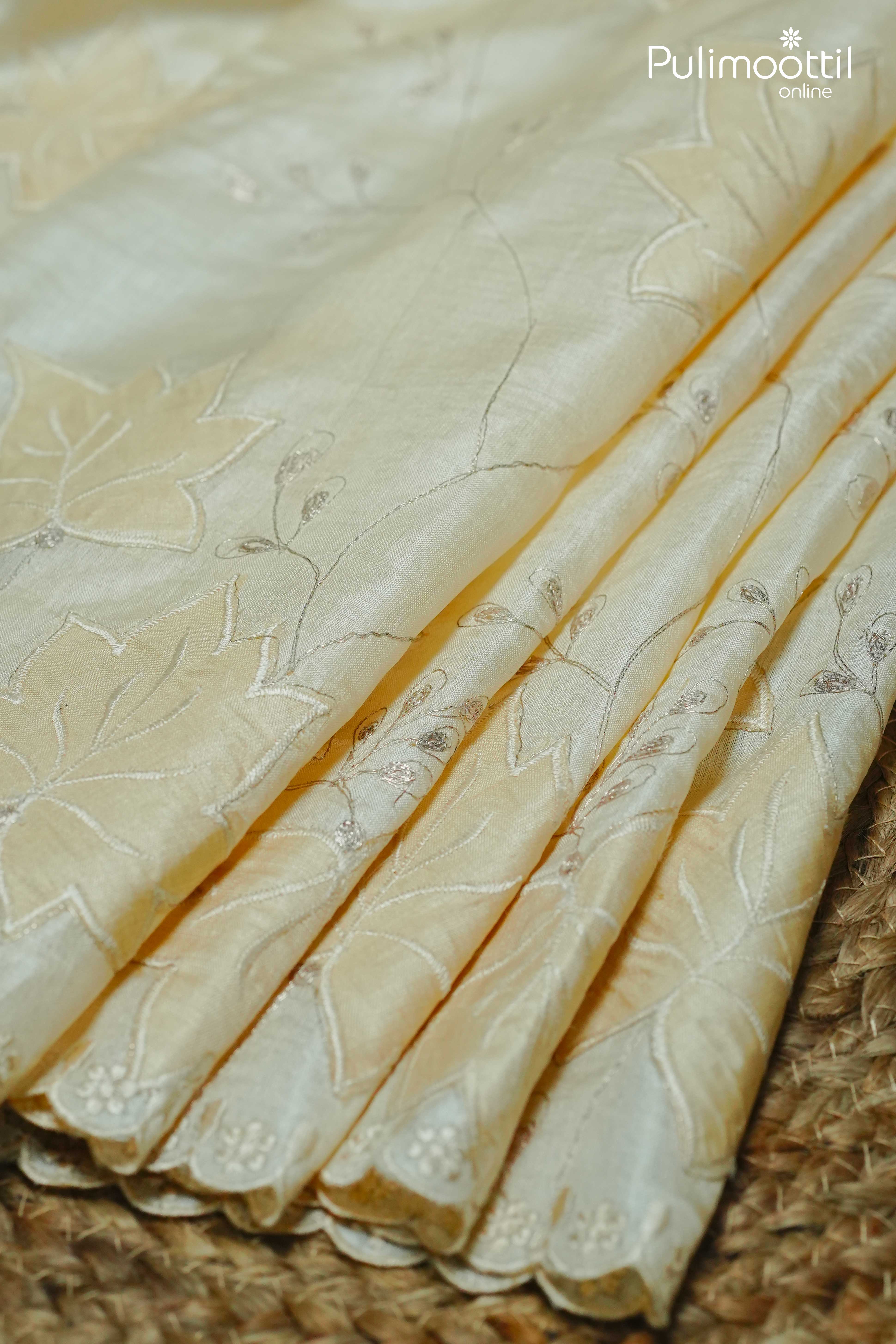 Close-up of folded off-white Tussar silk saree with floral applique embroidery and scallop border detailing, placed on a woven mat.