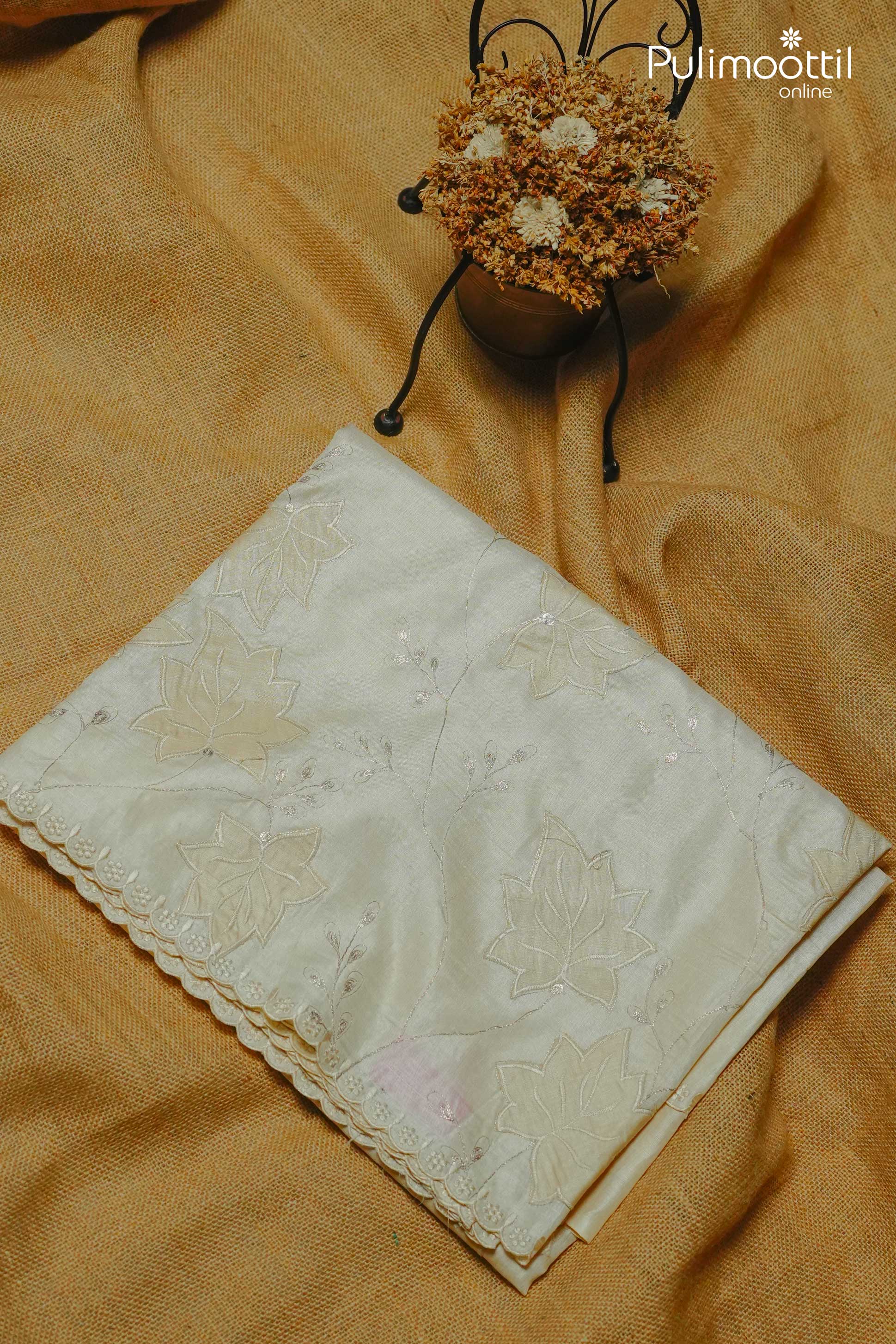 Folded off-white Tussar silk saree with floral appliqué work and scallop border placed on a brown textured fabric, alongside a pot of dried flowers.
