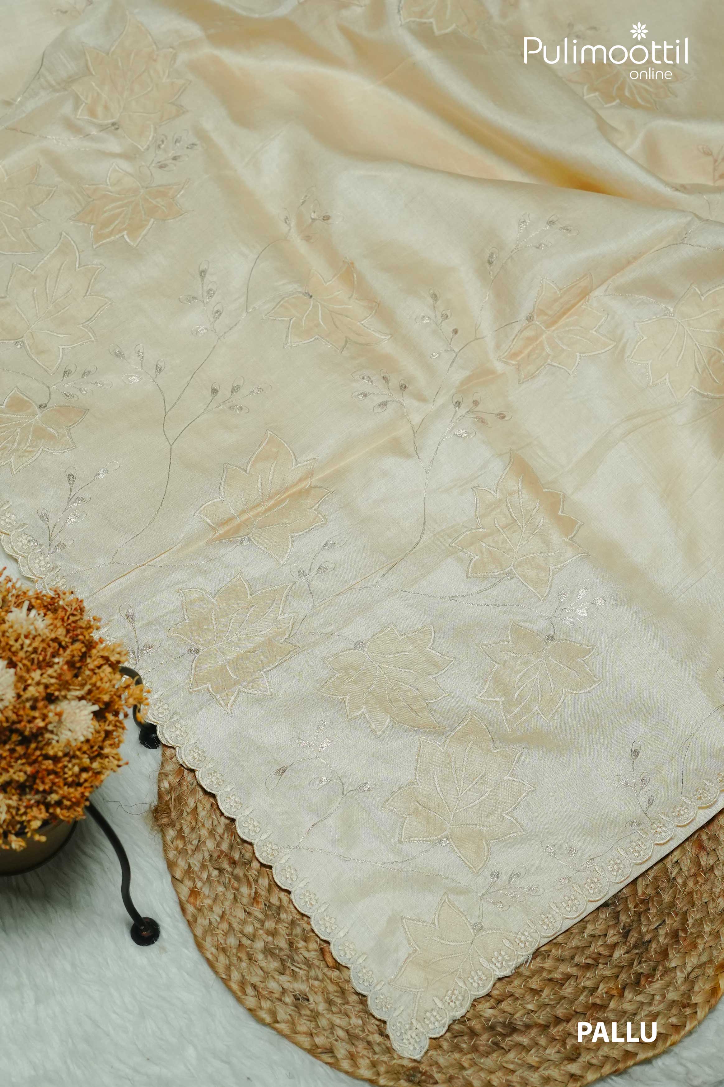 Close-up of the pallu of an off-white Tussar silk saree featuring intricate floral appliqué work and scallop border, displayed on a textured mat with dried flowers.